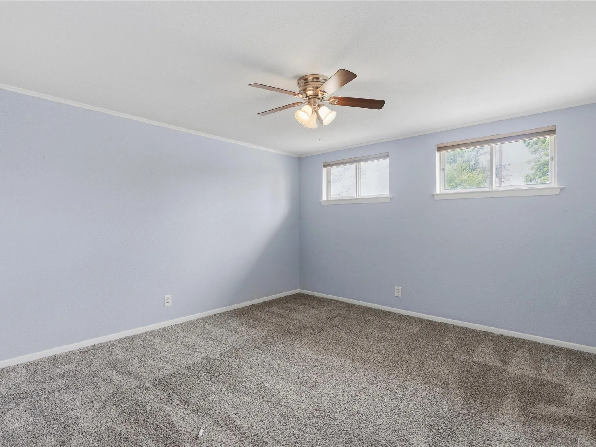Spacious interior room featuring light blue walls, grey patterned carpeting, a ceiling fan with light fixture, and two windows providing natural illumination
