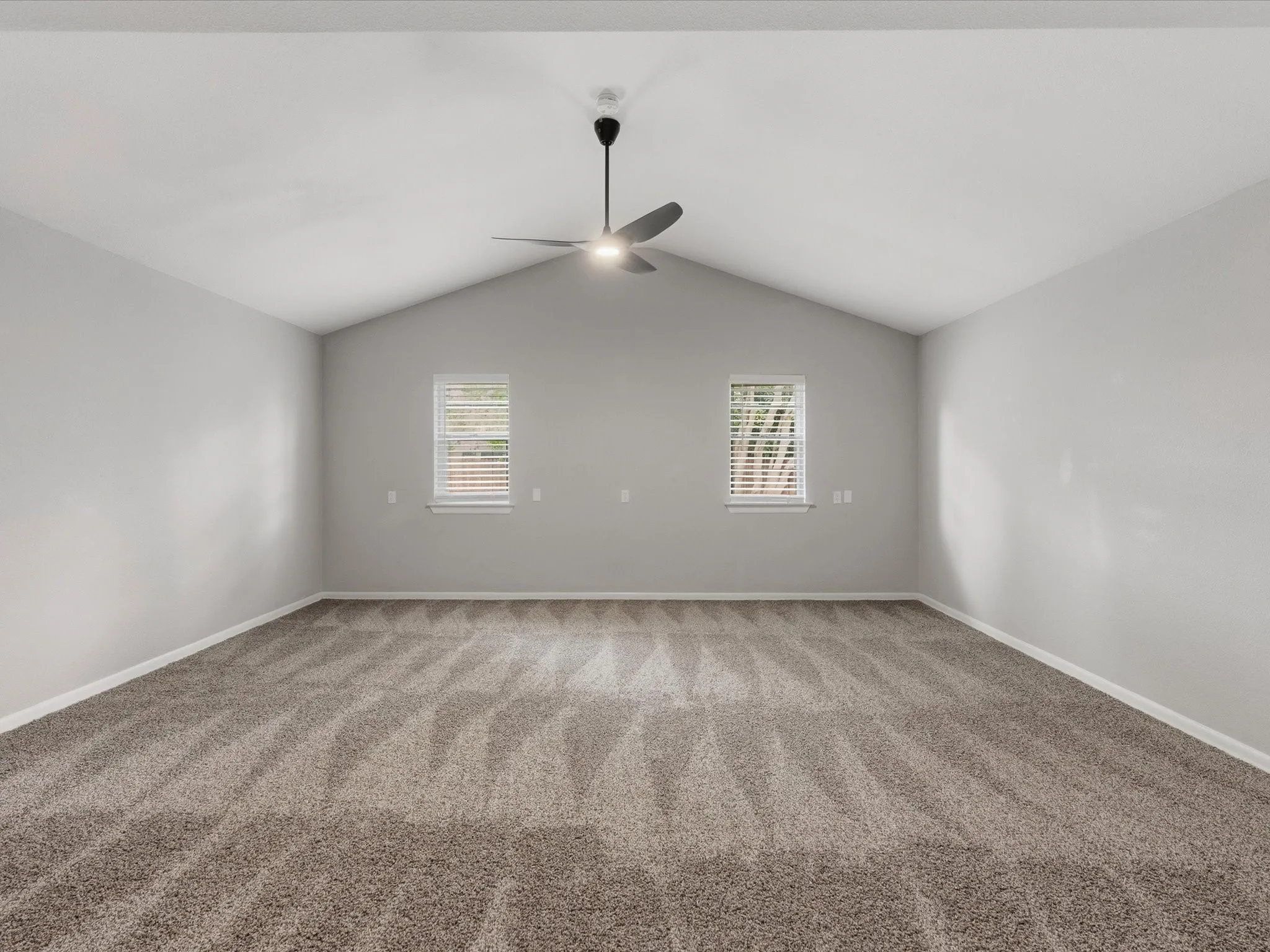 Spacious room featuring vaulted ceilings, a ceiling fan with integrated lighting, two windows with blinds, and textured carpeting