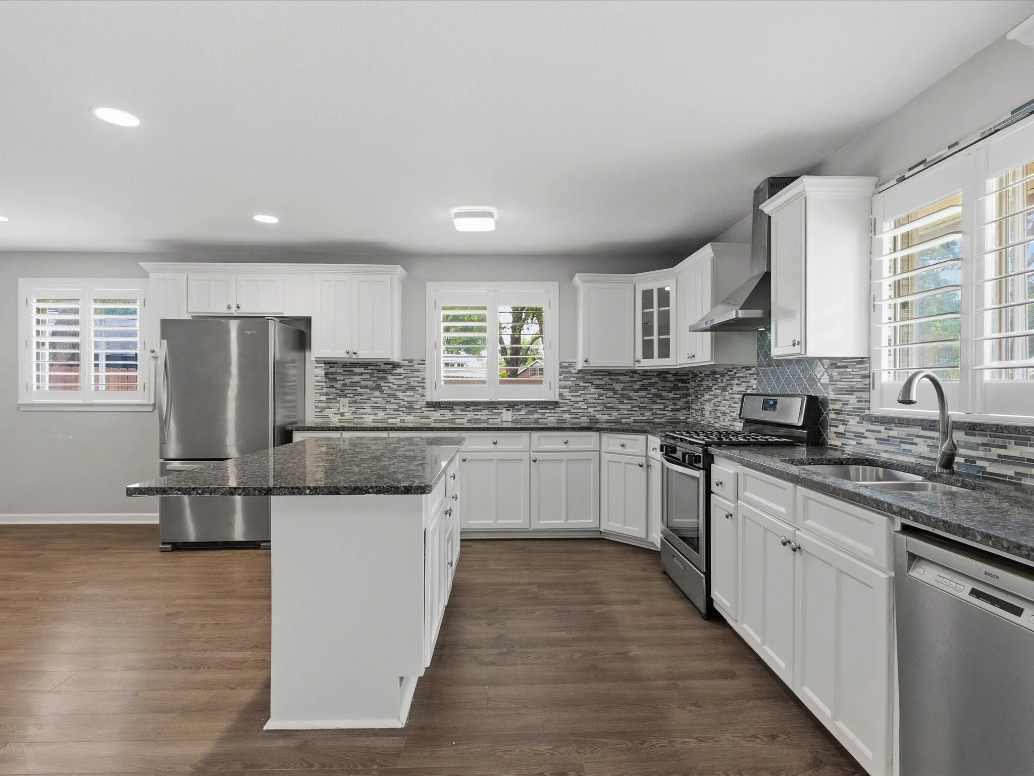 Spacious kitchen featuring an expansive island with stone countertop, white shaker cabinetry, stainless steel appliances, and wood-finish flooring