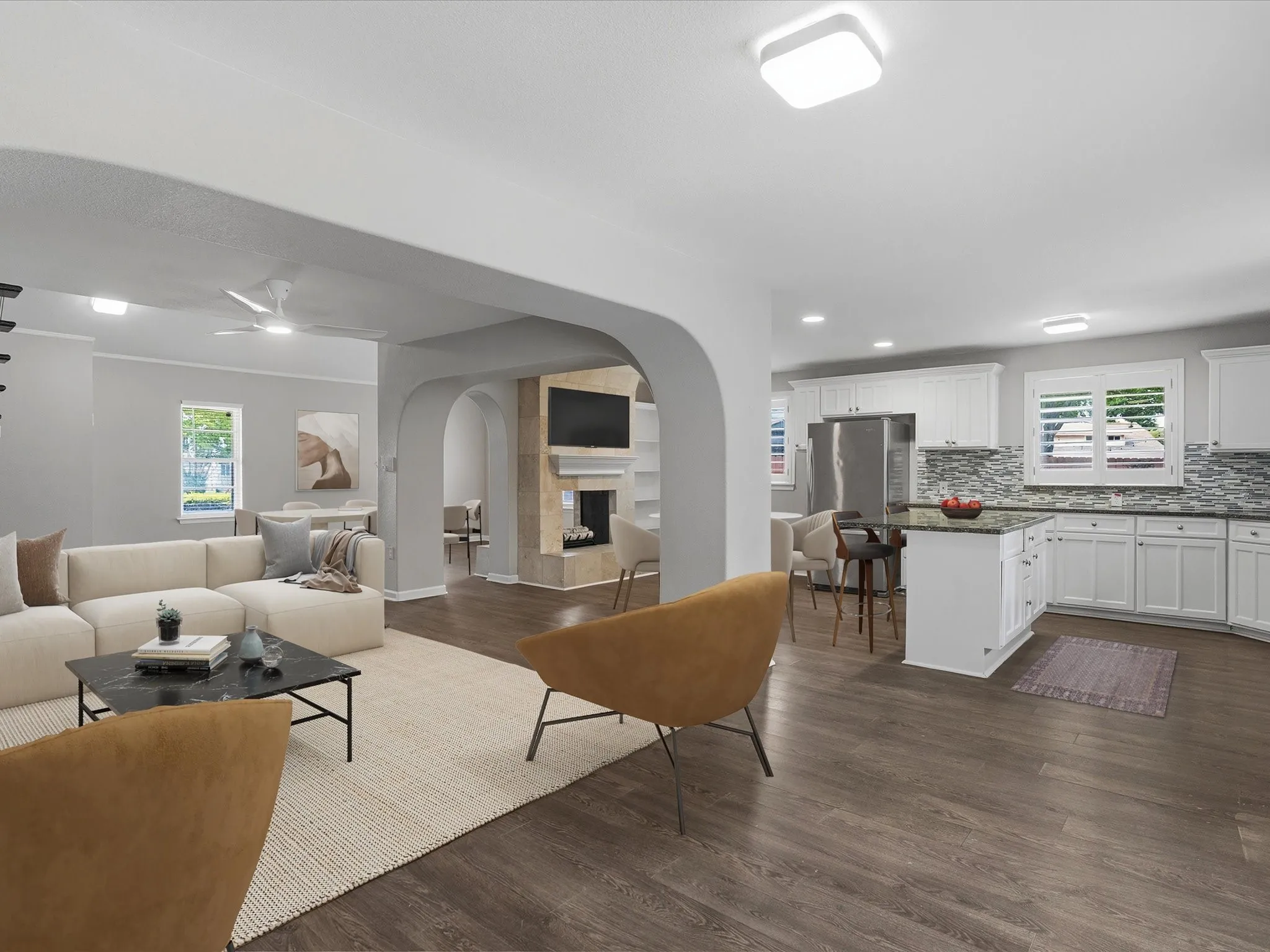 Open-concept living space featuring wood-finish flooring, a white kitchen with stone countertops and a mosaic tile backsplash, a built-in fireplace with a stone surround, and arched doorways