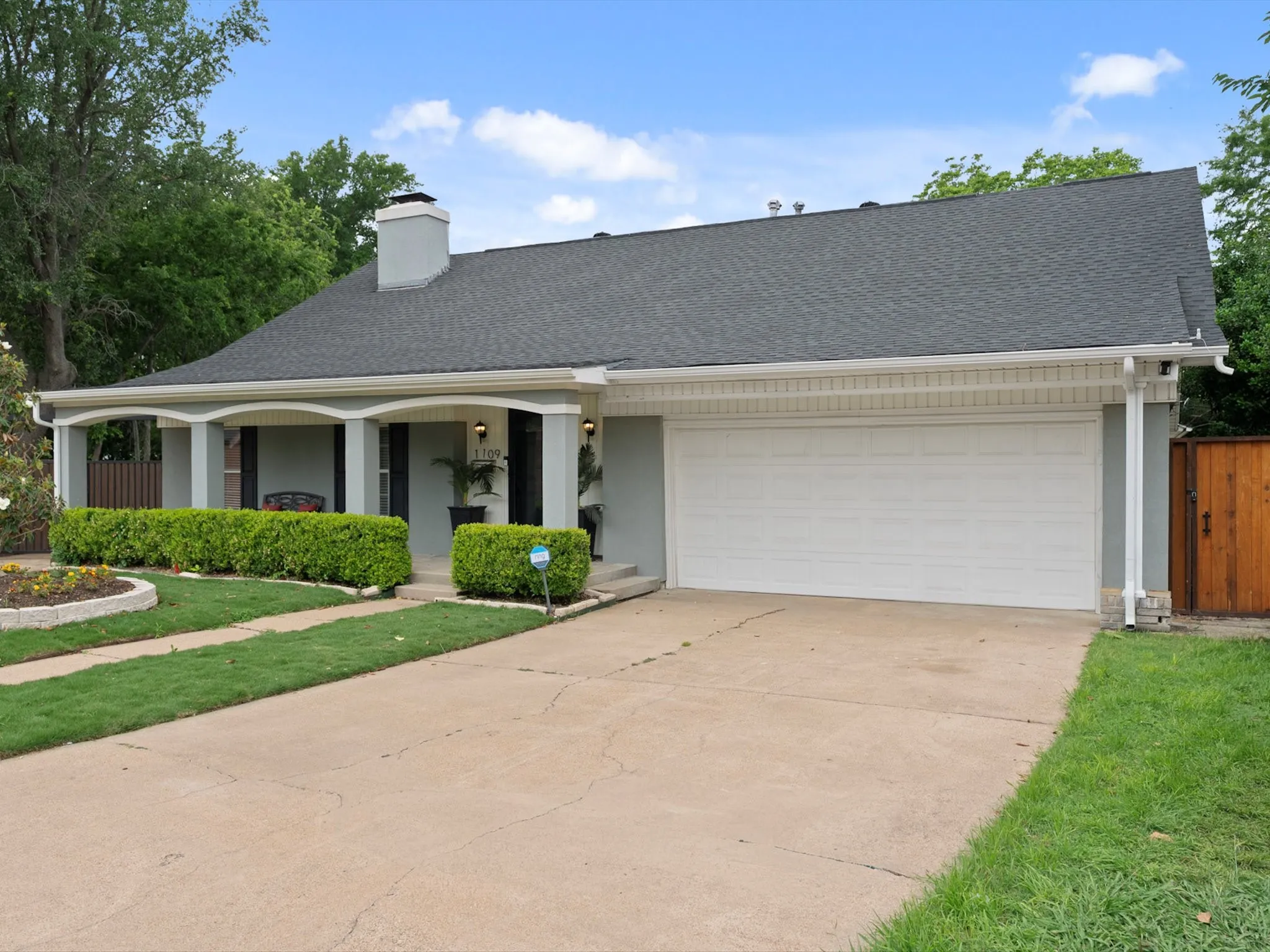 Exterior featuring a covered porch with arched accents, mature landscaping, and a two-car garage