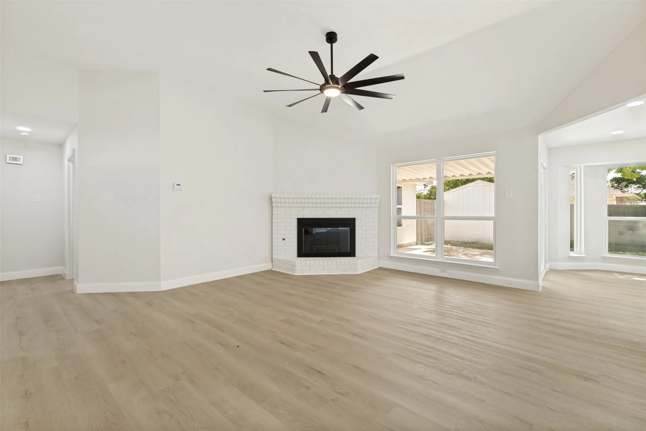 Spacious living area featuring vaulted ceilings, wood-finish flooring, a white brick fireplace, a modern ceiling fan, and multiple large windows