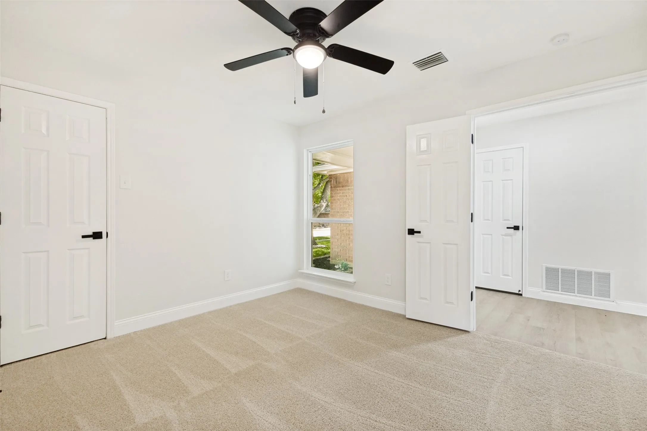 Spacious room featuring neutral paint, light-colored carpet, a ceiling fan with integrated lighting, a large window, and multiple panel doors with matte black hardware