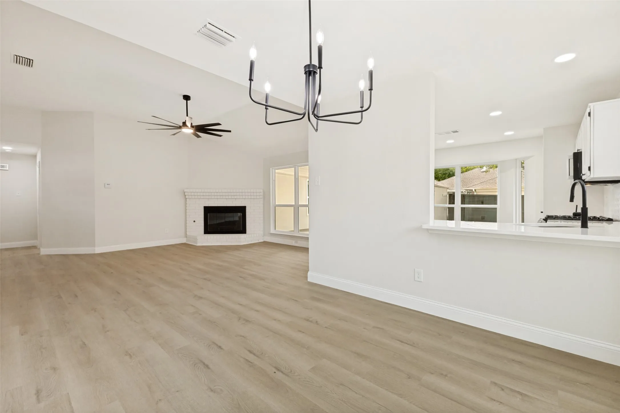 Open-concept living space featuring wood-finish flooring, a contemporary black chandelier, a corner fireplace with a white brick surround, vaulted ceilings, and a kitchen pass-through with a white countertop