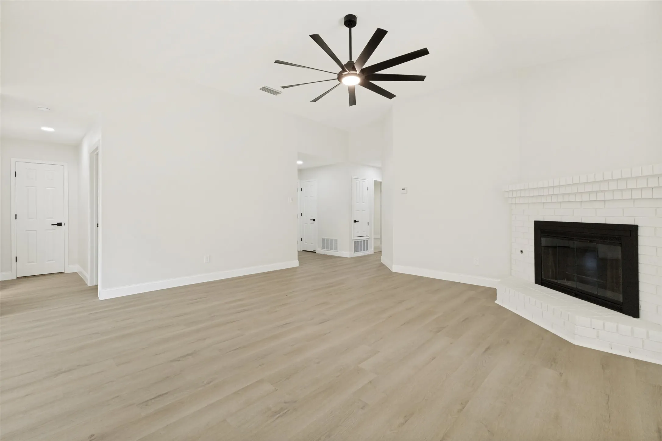 Spacious room featuring vaulted ceilings, wood-finish flooring, a modern ceiling fan with integrated lighting, and a white brick fireplace