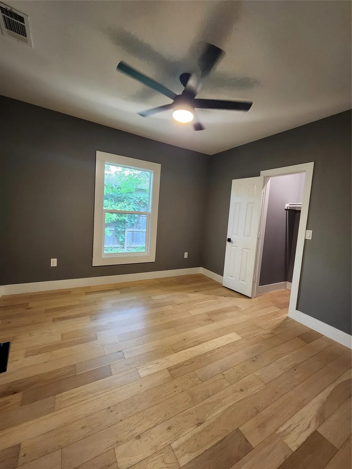 Spacious room featuring wood-finish flooring, a contemporary ceiling fan with integrated lighting, and a window with white trim