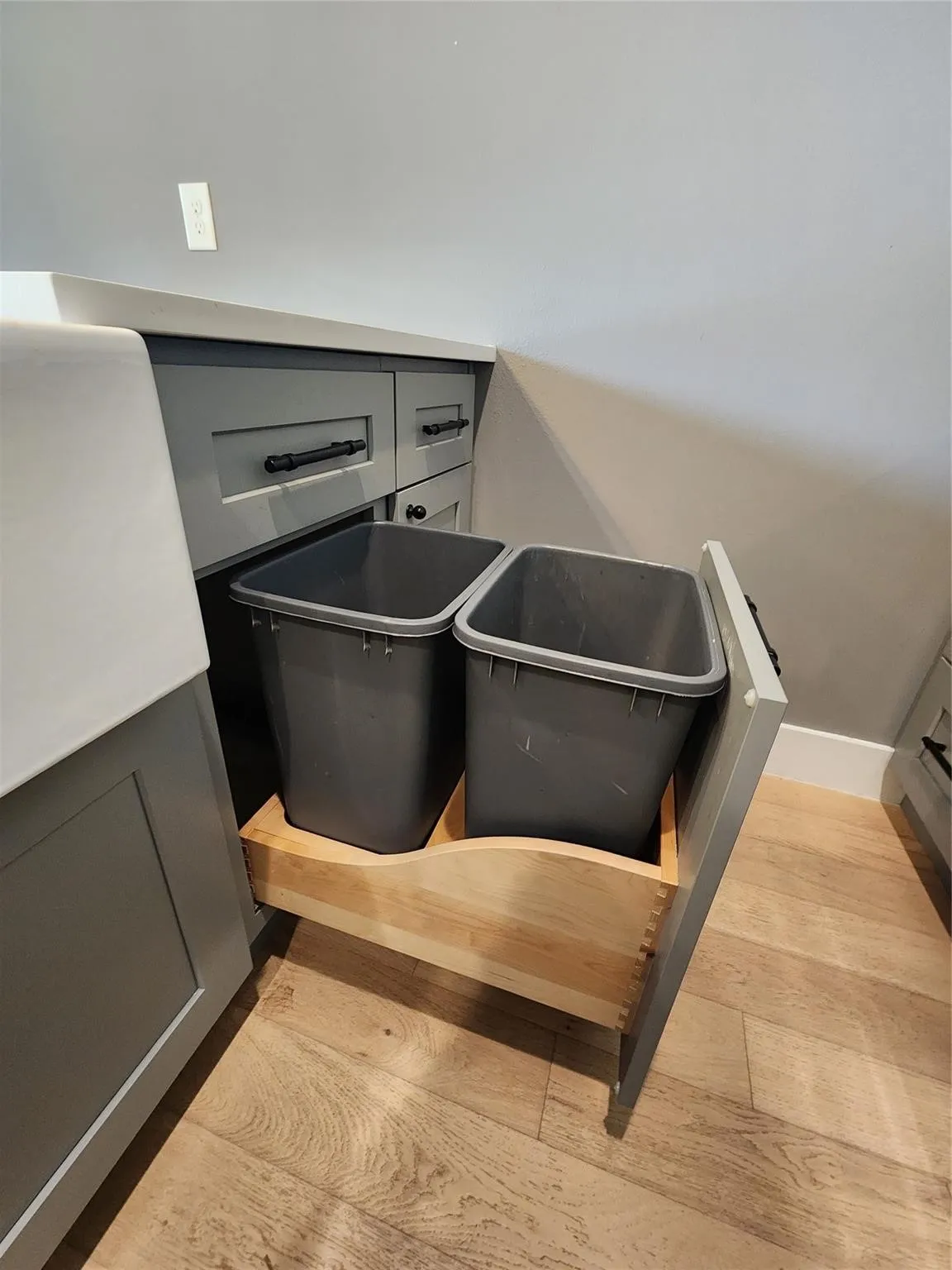 Integrated pull-out waste and recycling system within custom cabinetry, featuring wood-finish flooring, gray shaker-style cabinets with black hardware, and a solid surface countertop
