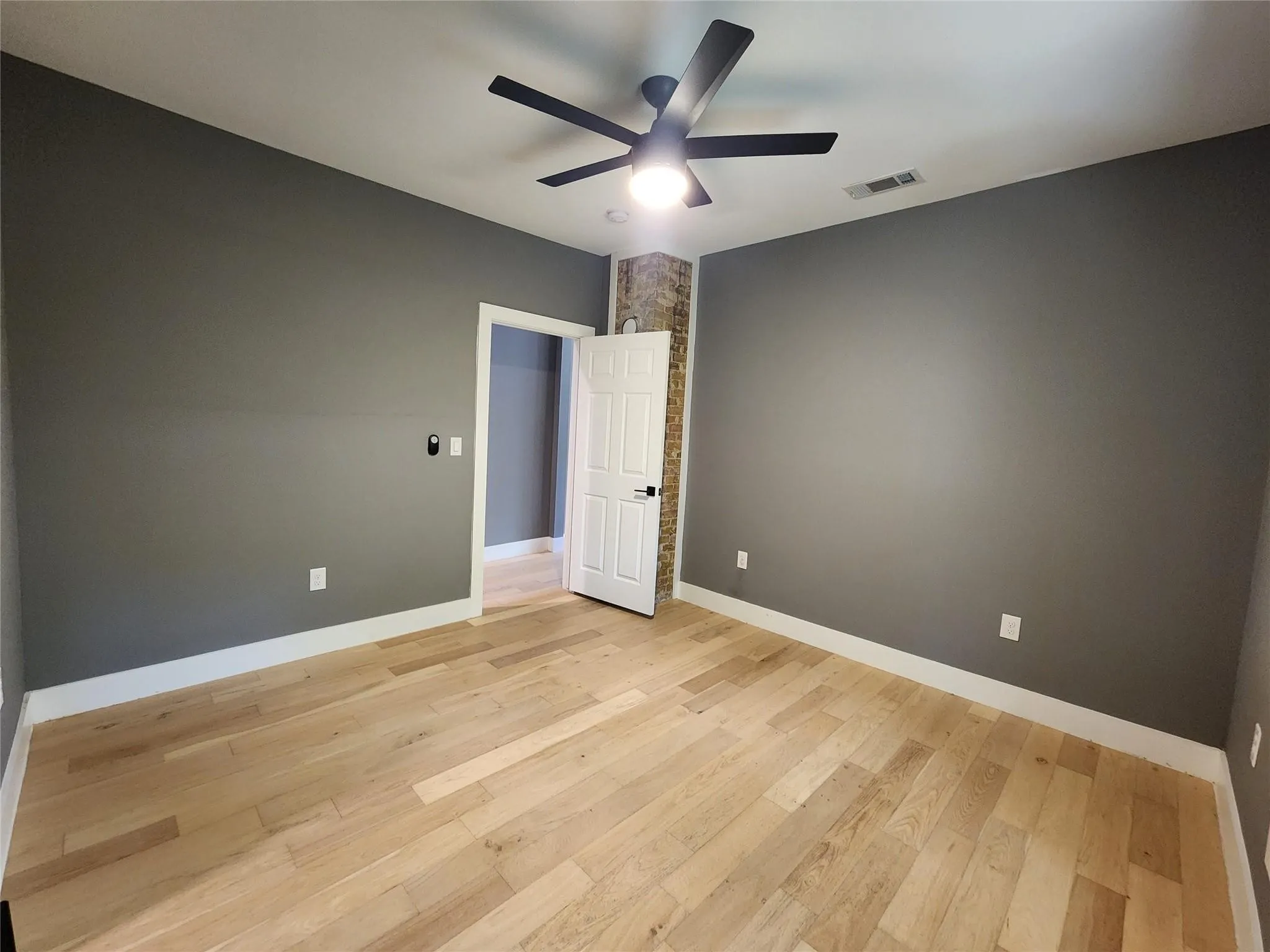 Spacious room featuring light wood-finish flooring, dark gray walls, white baseboards, a modern ceiling fan with integrated lighting, and an exposed brick accent wall