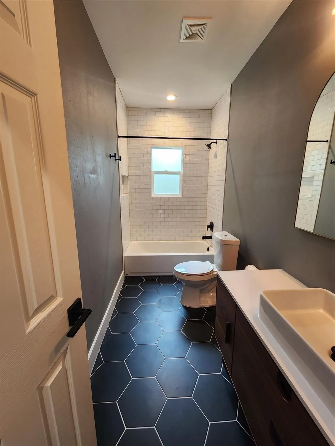 Contemporary bathroom featuring dark hexagonal floor tiling, a white subway-tiled shower surround, a white tub, and a dark wood-finish vanity with a white countertop and vessel sink