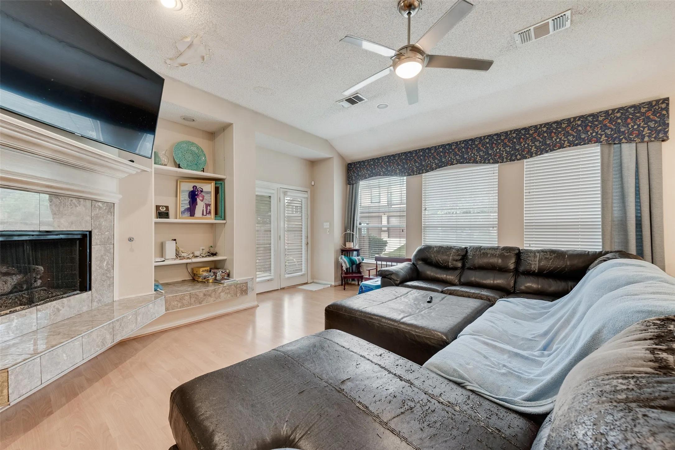 Living area featuring light wood-finish flooring, a tile fireplace with mantel, built-in shelving, a ceiling fan, and multiple windows