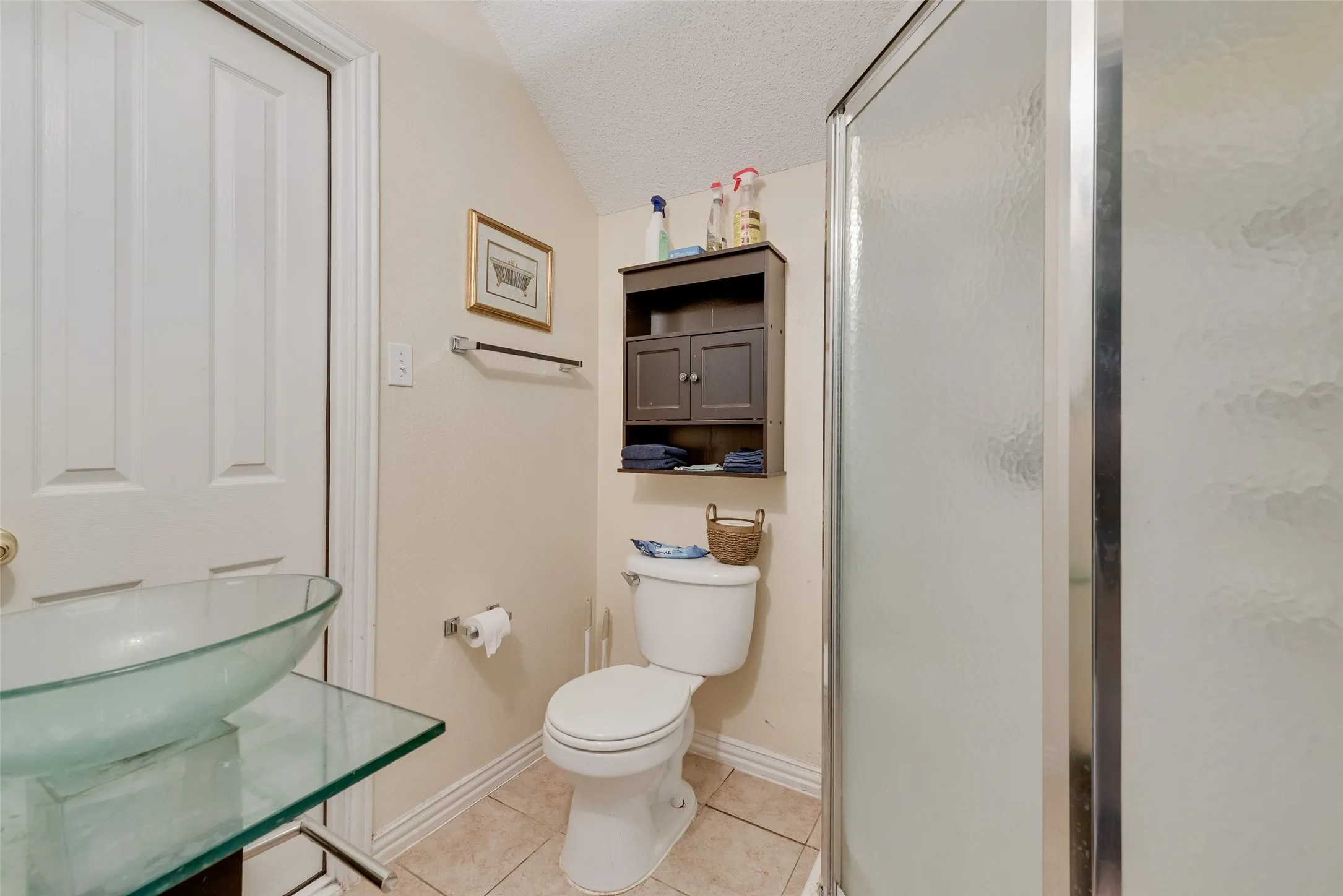 Bathroom featuring a glass vessel sink, glass countertop, a toilet, a dark wood-finish wall cabinet, and a shower with a frosted glass enclosure