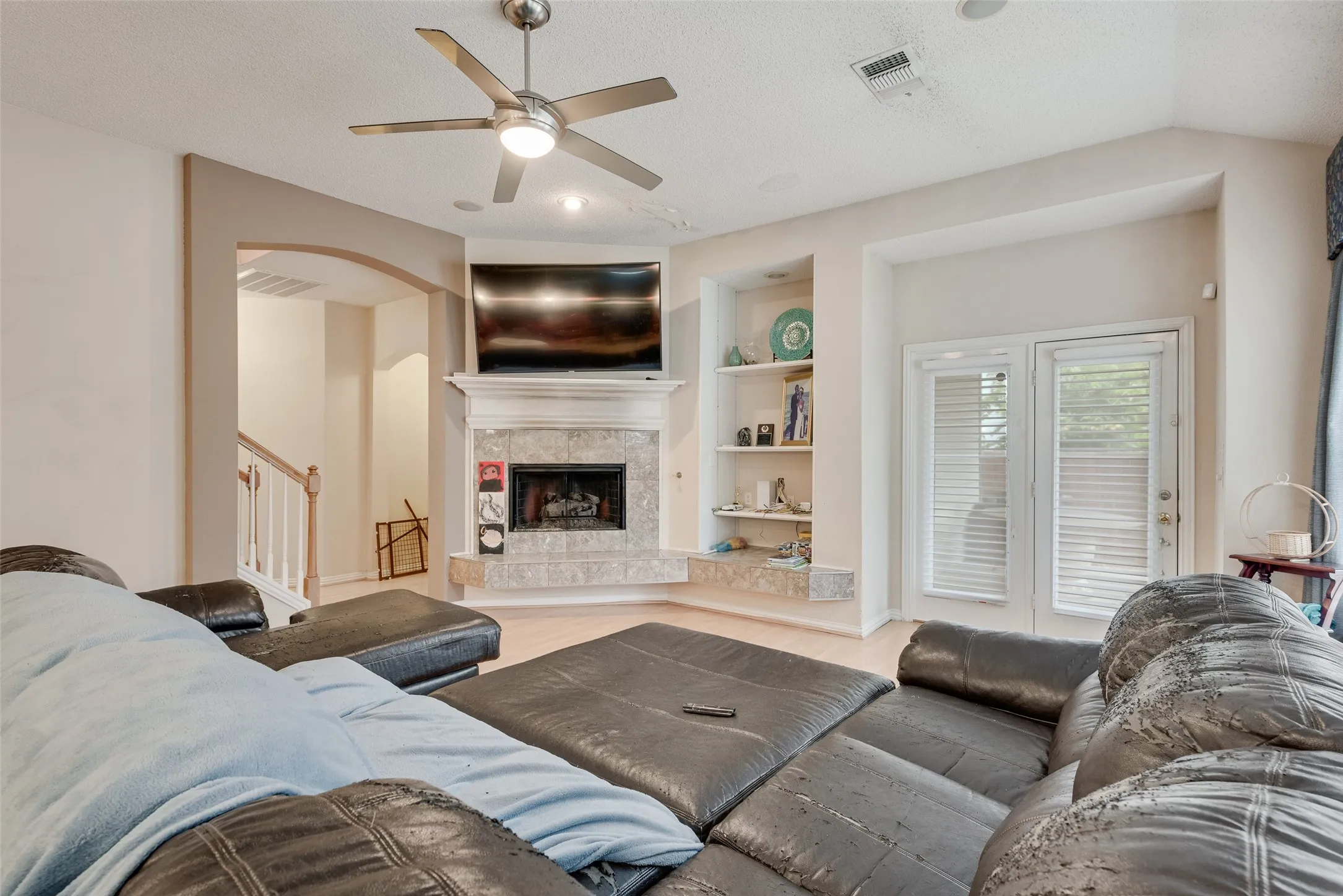 Living area featuring a corner fireplace with a tiled surround and mantel, built-in shelving, a ceiling fan with integrated lighting, and light wood-finish flooring