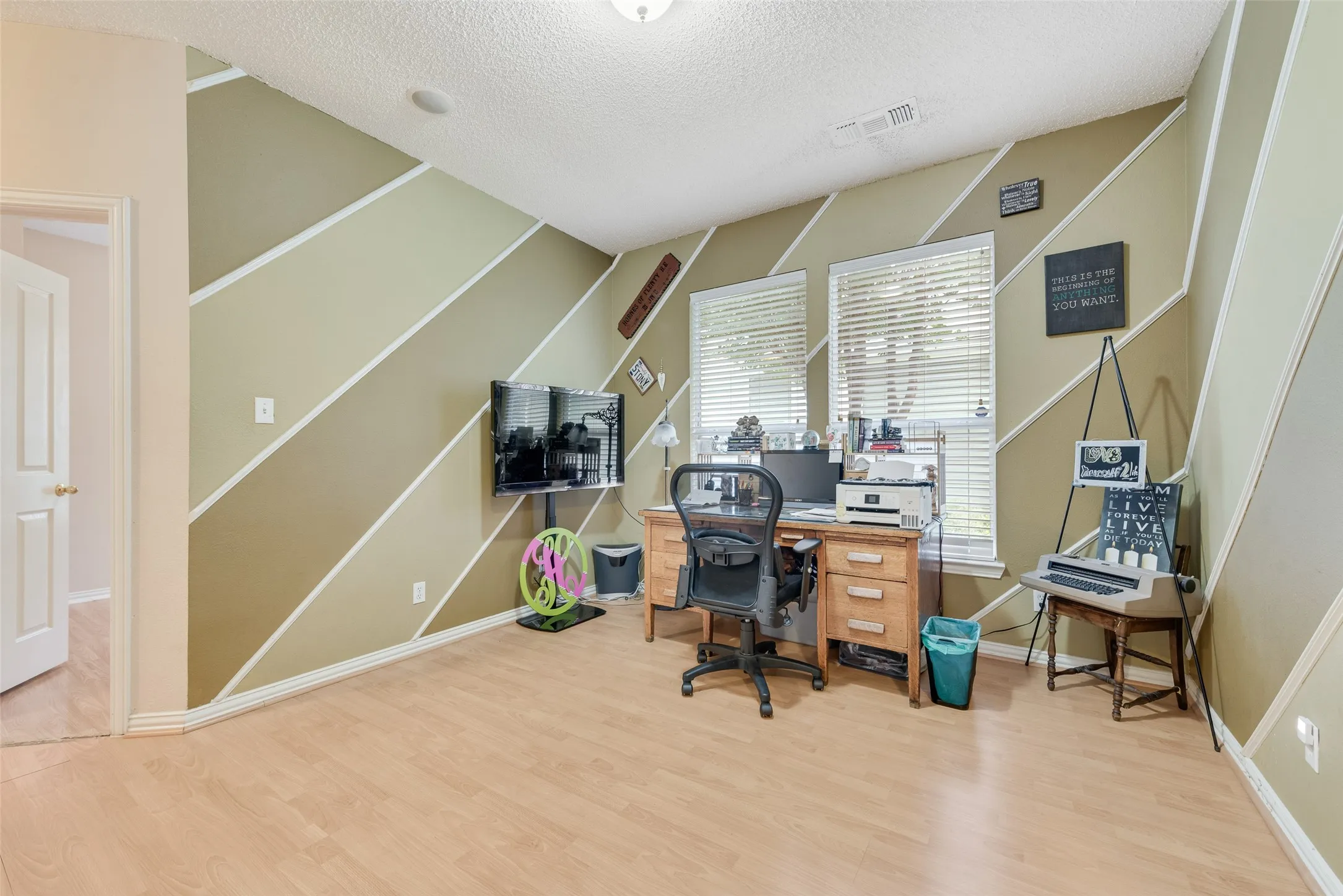 Spacious room featuring wood-finish flooring, two large windows with blinds, a wall-mounted television, an open doorway, and a ceiling light fixture