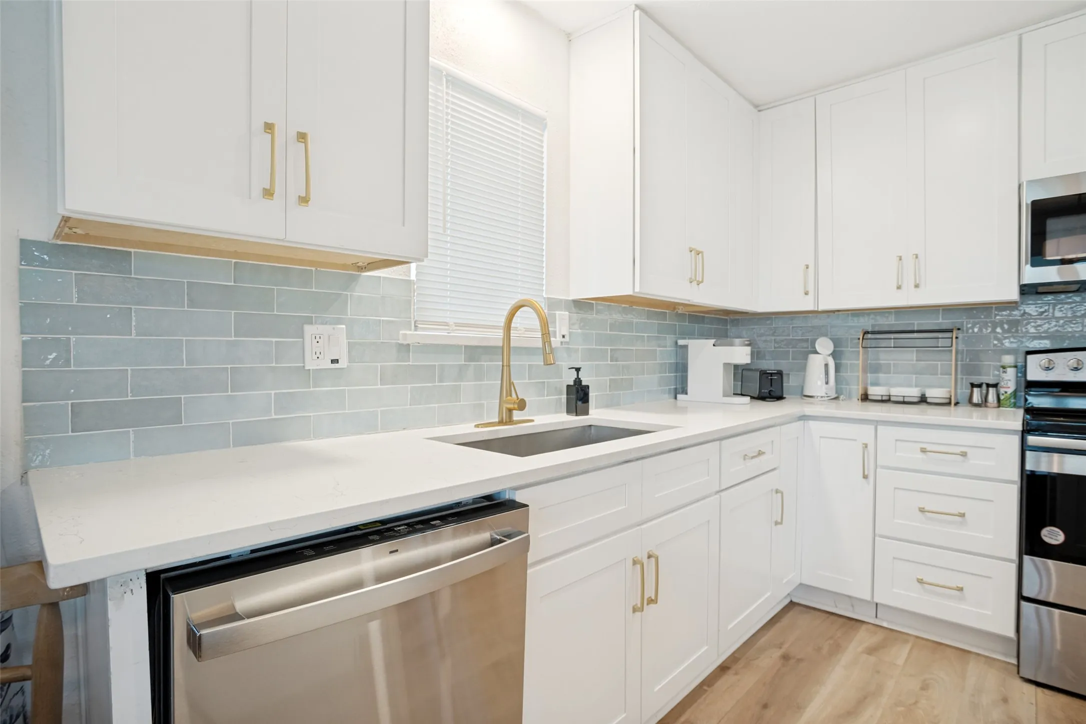 Modern kitchen featuring white shaker cabinetry, light-toned countertops, a stainless steel dishwasher, a gold-finish faucet, and light blue subway tile backsplash