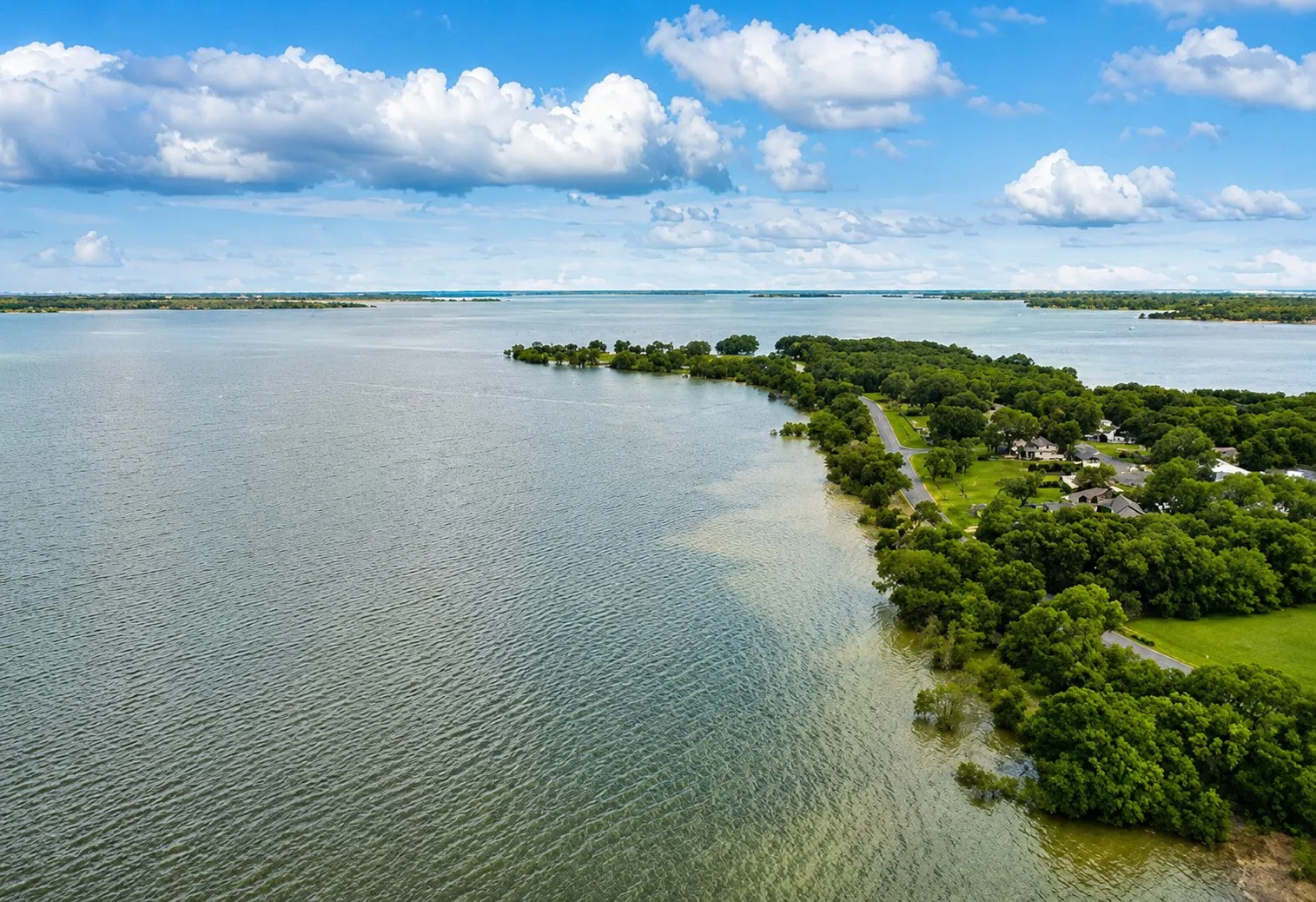 Expansive waterfront setting featuring a large body of water, lush green tree lines, and a residential area with multiple homes