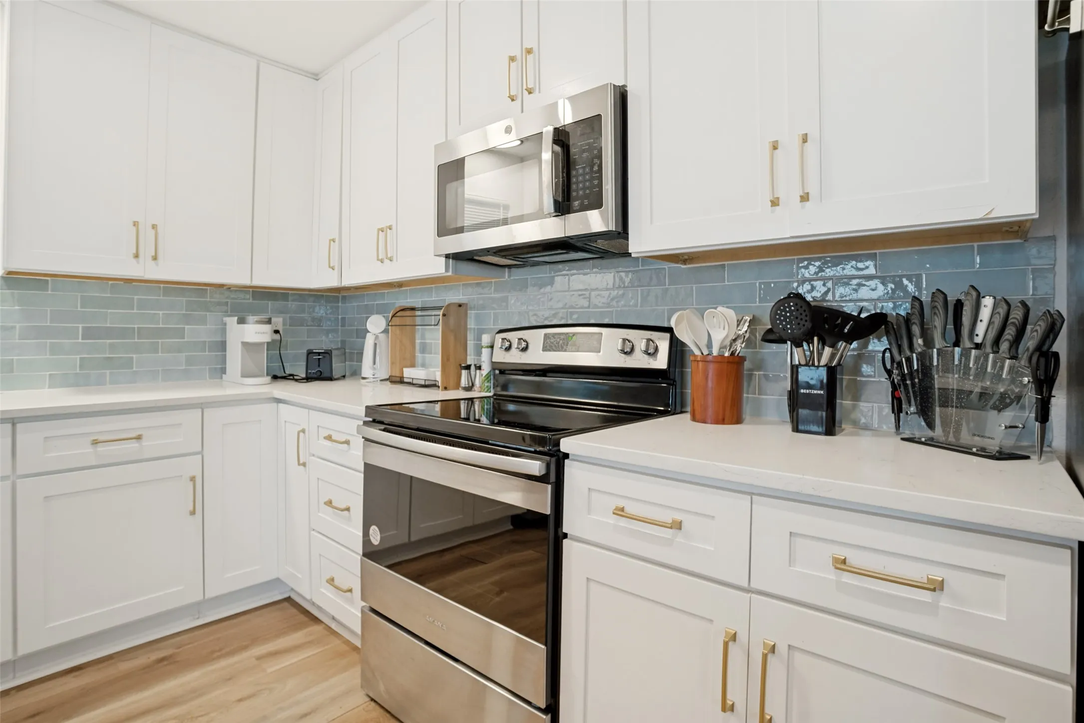 Modern kitchen featuring white cabinetry with gold hardware, light-colored countertops, a stylish blue subway tile backsplash, stainless steel appliances, and wood-finish flooring
