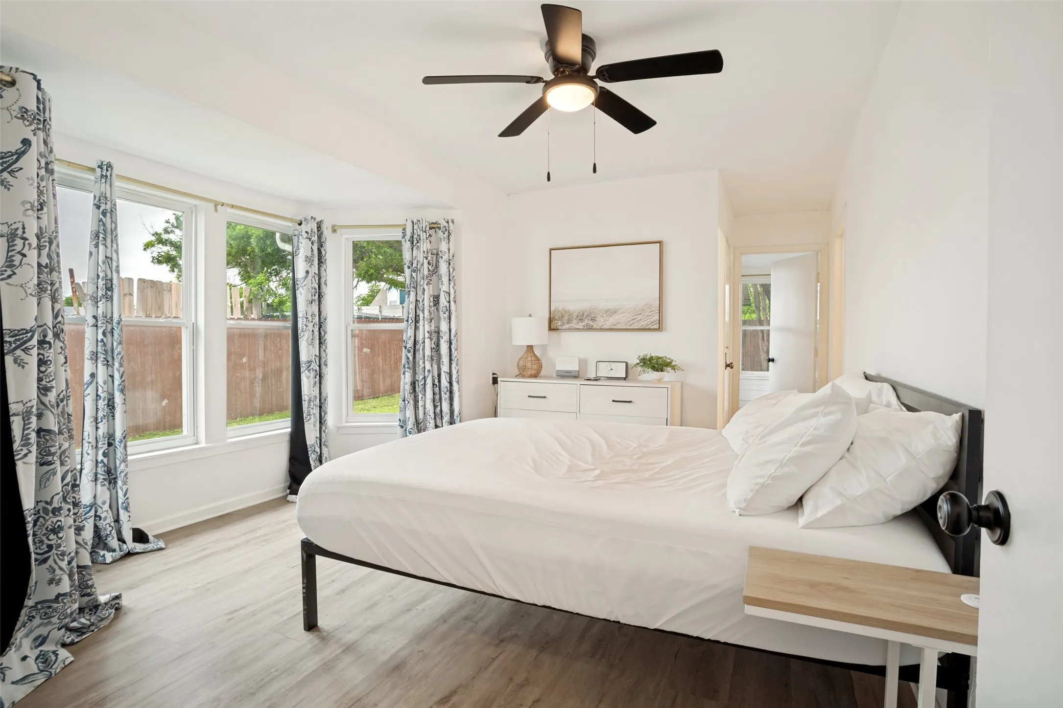 Light-filled room featuring a bay window with multiple panes, wood-finish flooring, a ceiling fan with integrated lighting, and a white dresser with black hardware