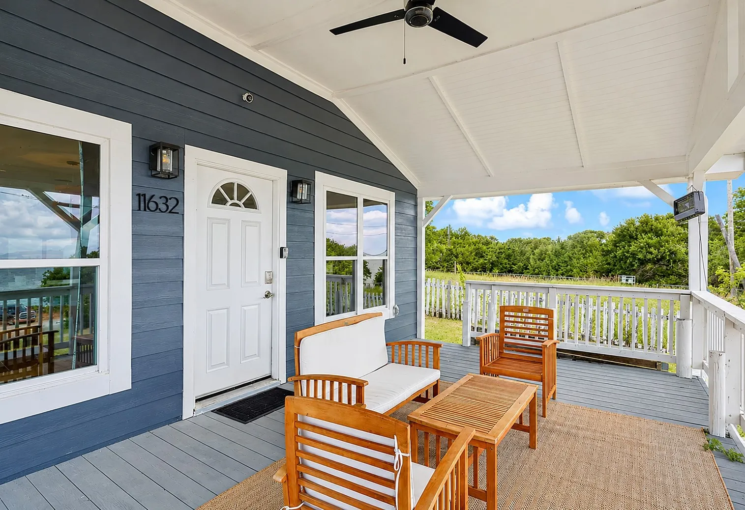 Covered front porch featuring a dark blue siding exterior with white trim, a white paneled door with a sunburst window, and a ceiling fan