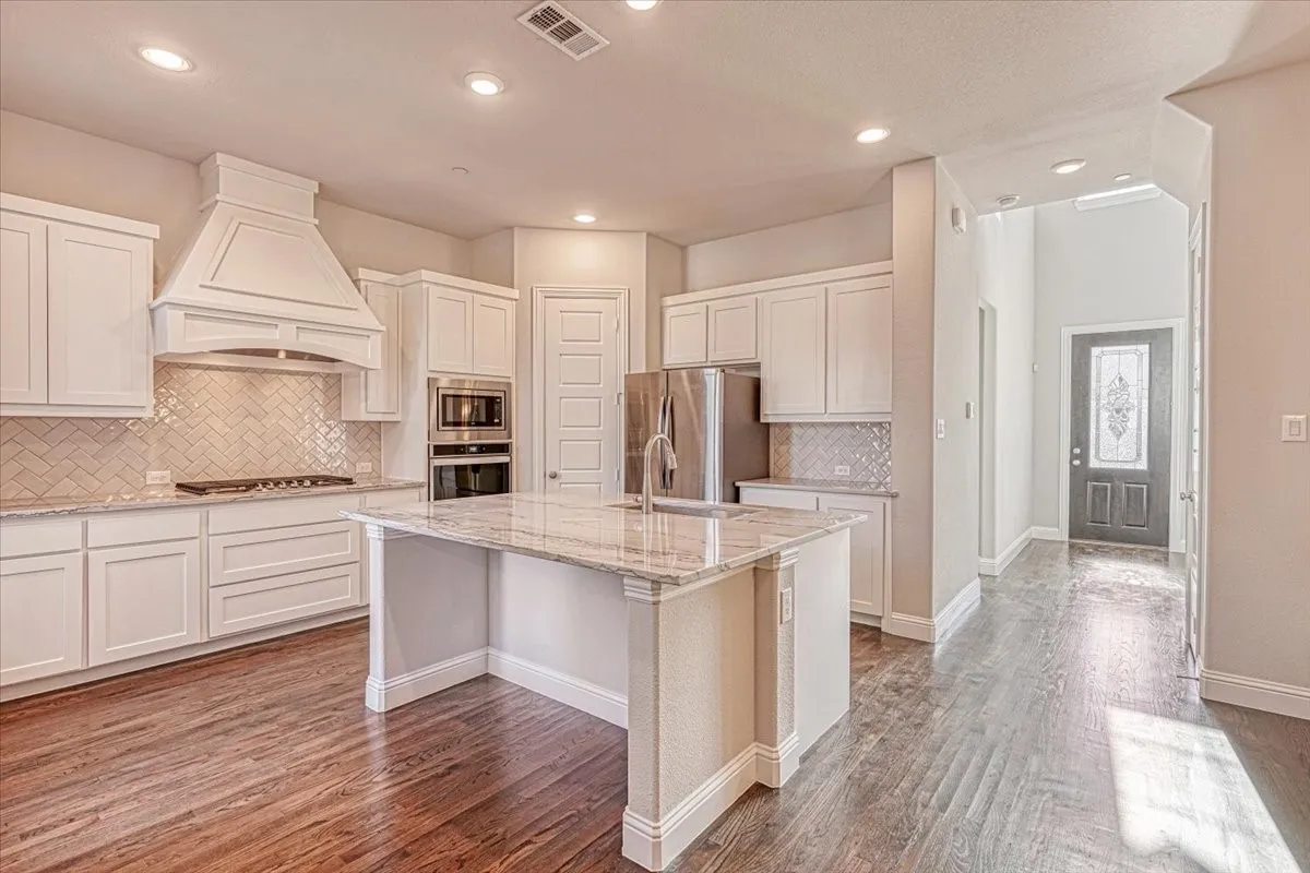 Kitchen featuring a center island with sink, wood-type flooring, premium range hood, backsplash, and appliances with stainless steel finishes