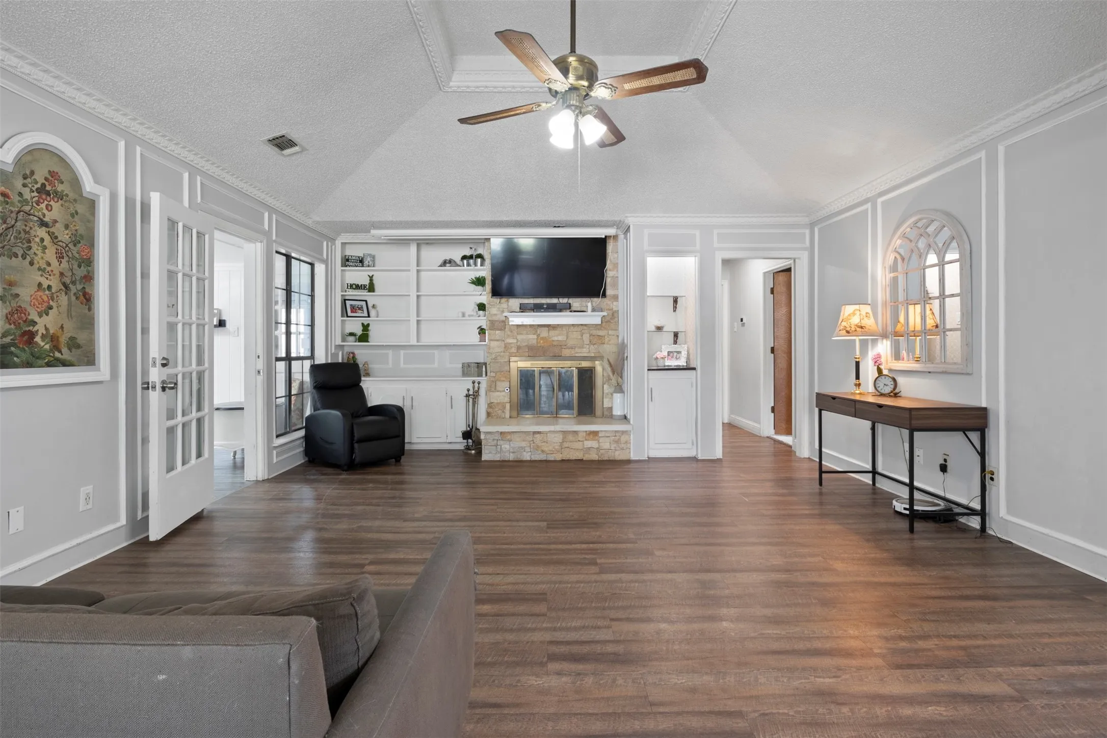 Vaulted ceiling living area featuring wood-finish flooring, a stone fireplace with built-in shelving, French doors, and crown molding details
