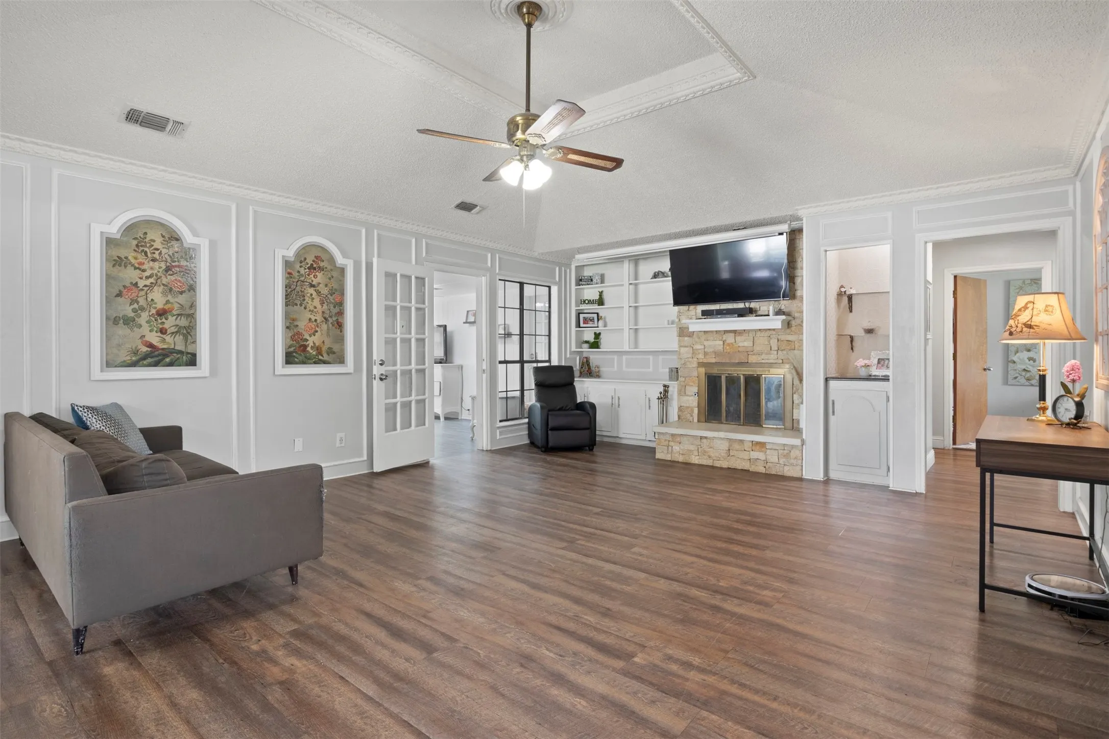 Spacious living area featuring wood-finish flooring, a stone fireplace with built-in shelving, vaulted ceiling with a fan, and white French doors
