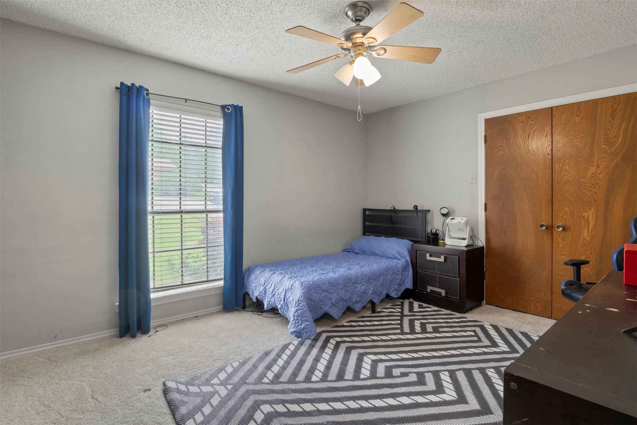 Carpeted room featuring a double-paned window with horizontal blinds, a ceiling fan with integrated lighting, and light wood-finish bi-fold closet doors