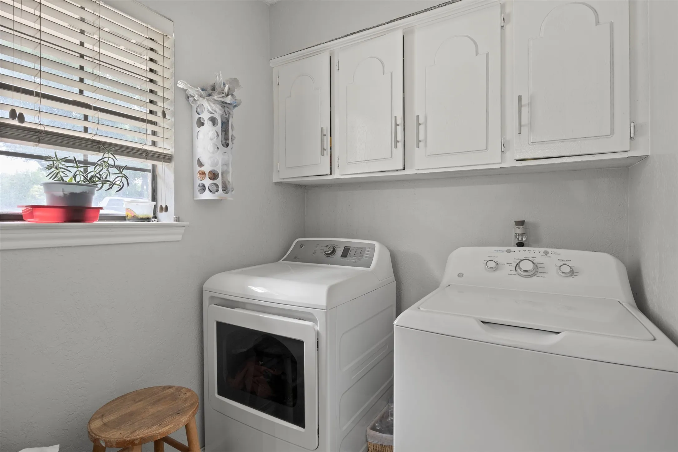 Dedicated laundry area featuring overhead white cabinetry with arched panel doors, a window with blinds, and light gray wall paint