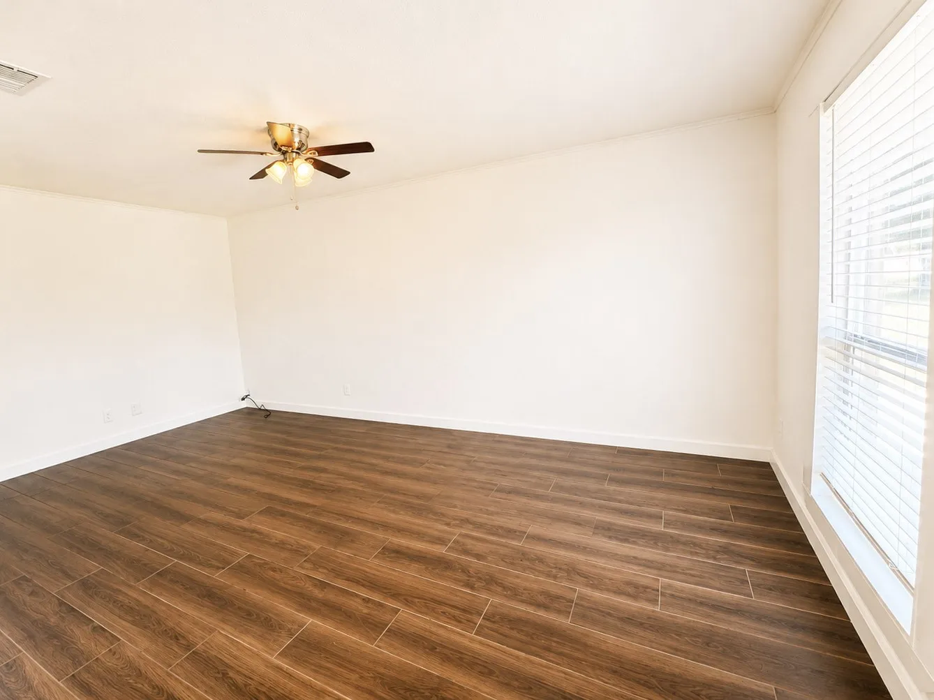 Spacious room featuring wood-finish flooring, light-toned walls, a ceiling fan with integrated lighting, white baseboards, and a window with blinds