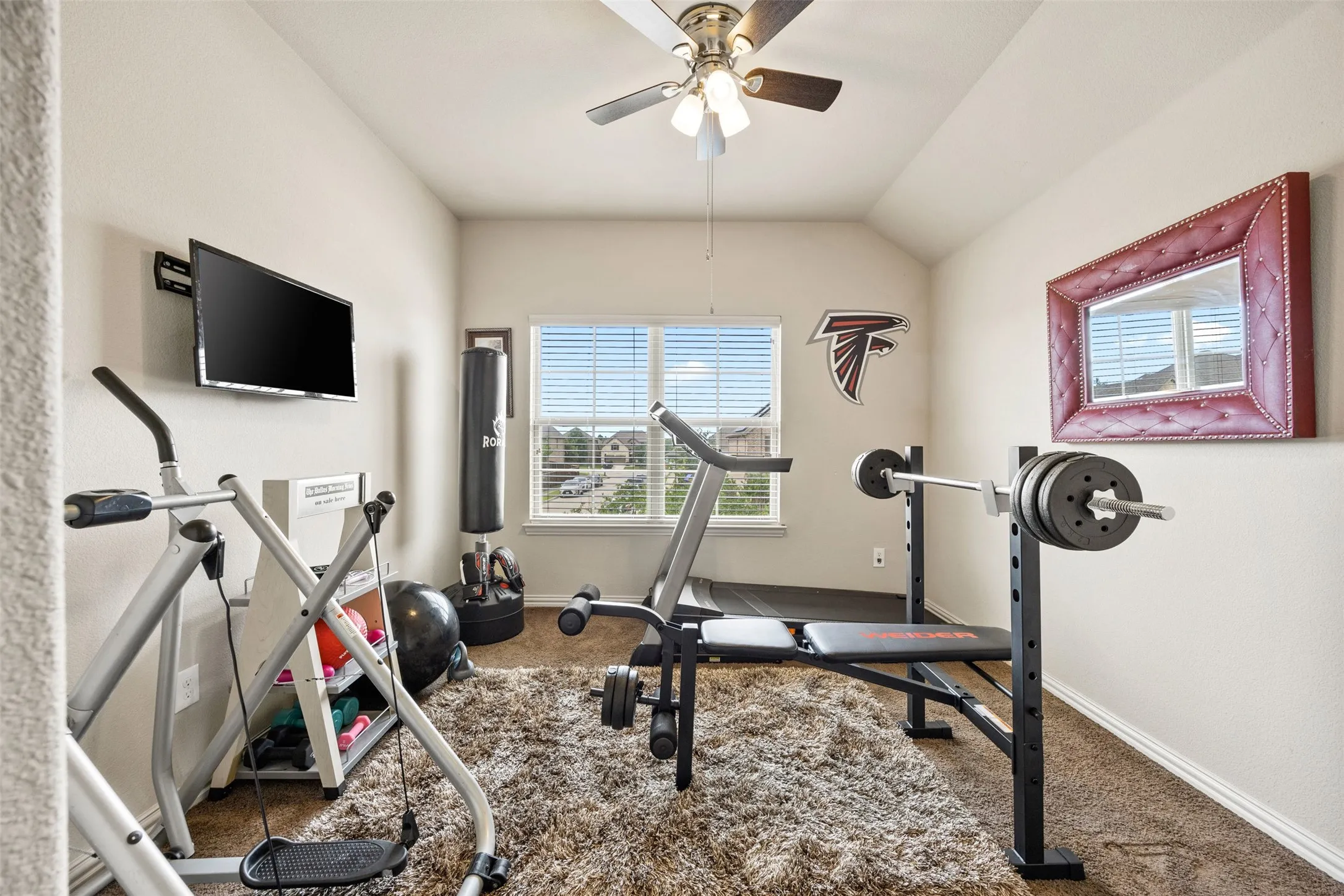 Versatile bonus room featuring a window with blinds, ceiling fan with integrated lighting, wall-mounted television bracket, and recessed wall mirror
