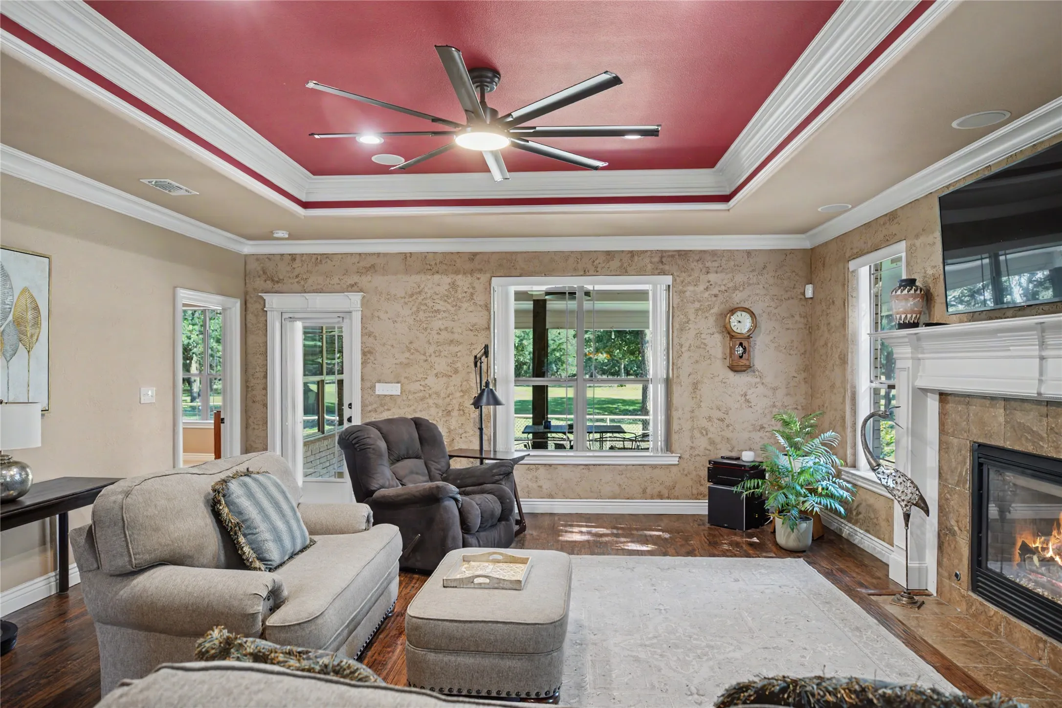 Spacious living area featuring a tray ceiling with a modern ceiling fan, wood-finish flooring, a built-in fireplace with a stone surround, and multiple windows allowing natural light