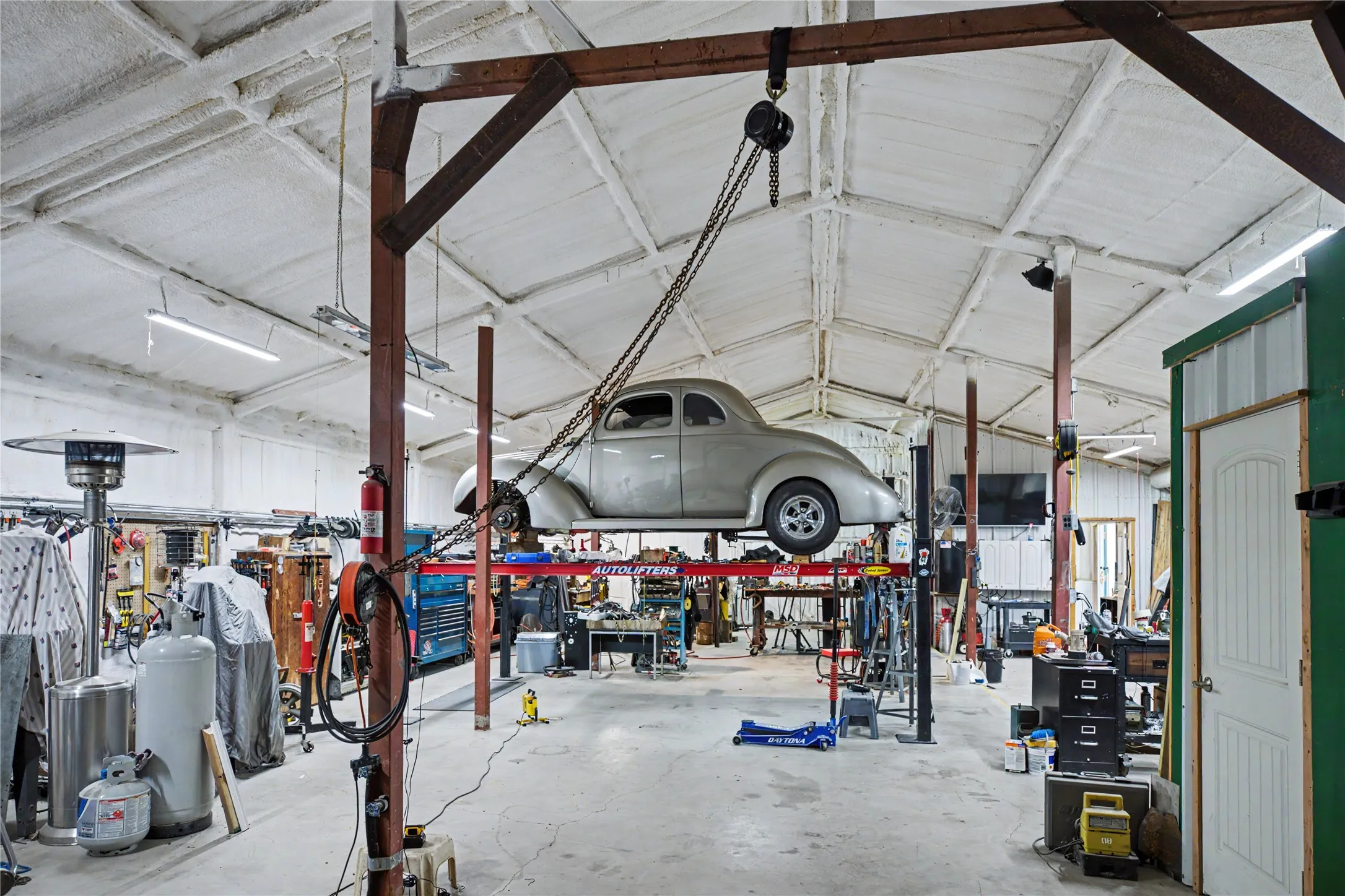 Spacious garage workshop featuring a vehicle lift, vaulted ceiling with exposed beams, fluorescent lighting, and a fire extinguisher