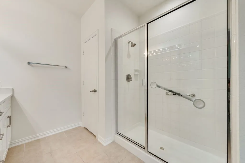 Bathroom featuring a walk-in shower with clear glass enclosure and grab bars, neutral-toned square tile flooring, a white vanity with a light-colored countertop, and a brushed nickel towel bar