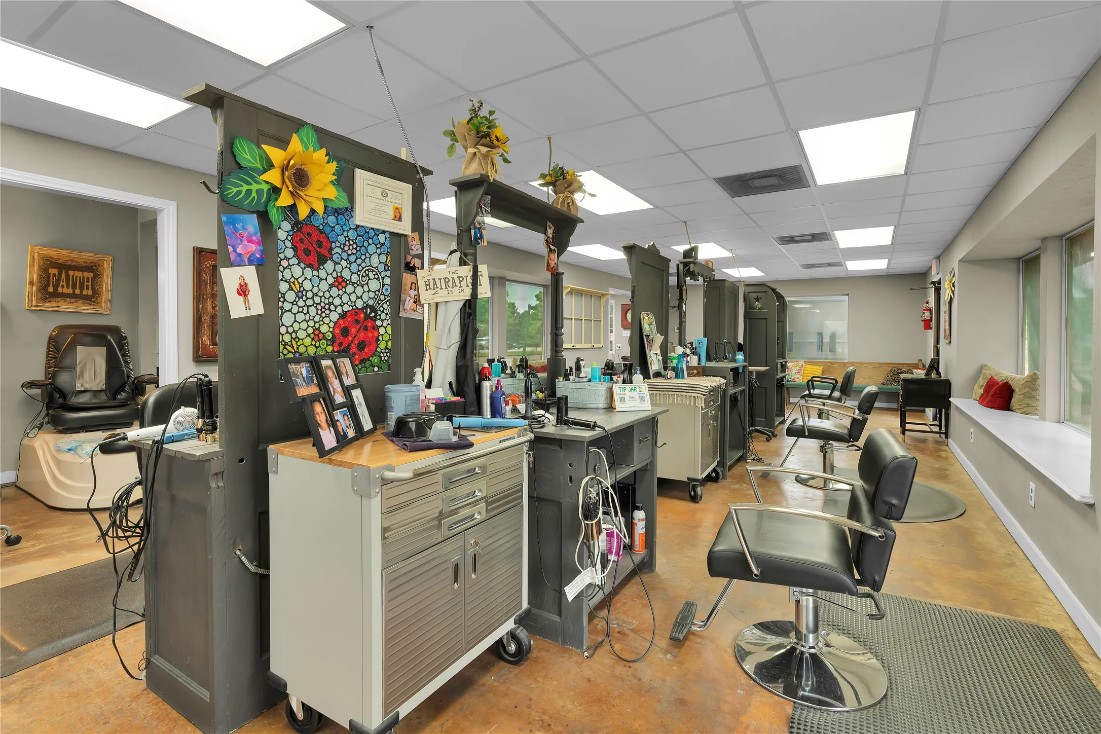 Commercial interior featuring a drop ceiling with recessed lighting, multiple workstations with partitioned mirrors, and a window bench with exterior views