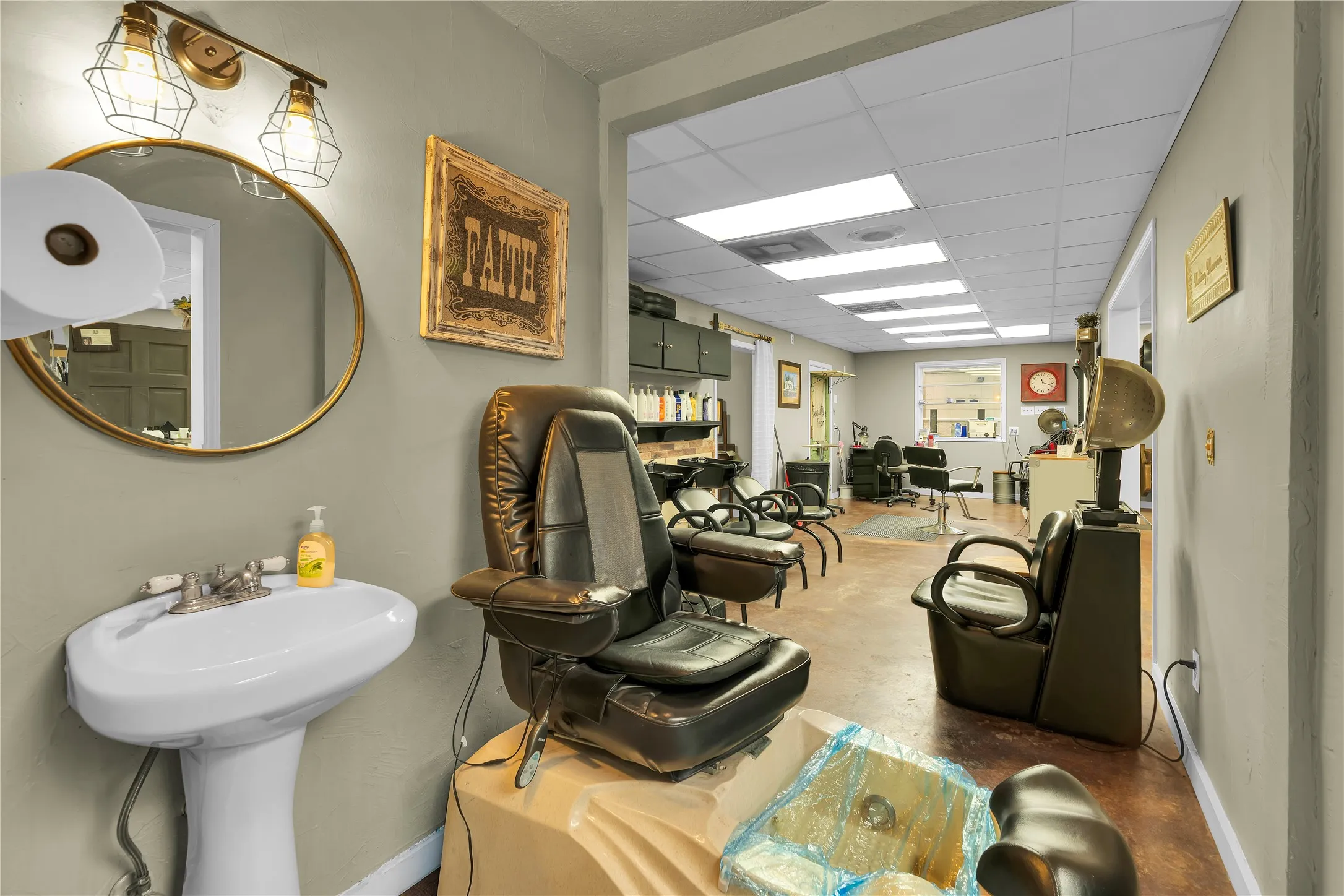 Brightly lit commercial space featuring a drop ceiling with recessed lighting and a pedestal sink with a round gold-framed mirror