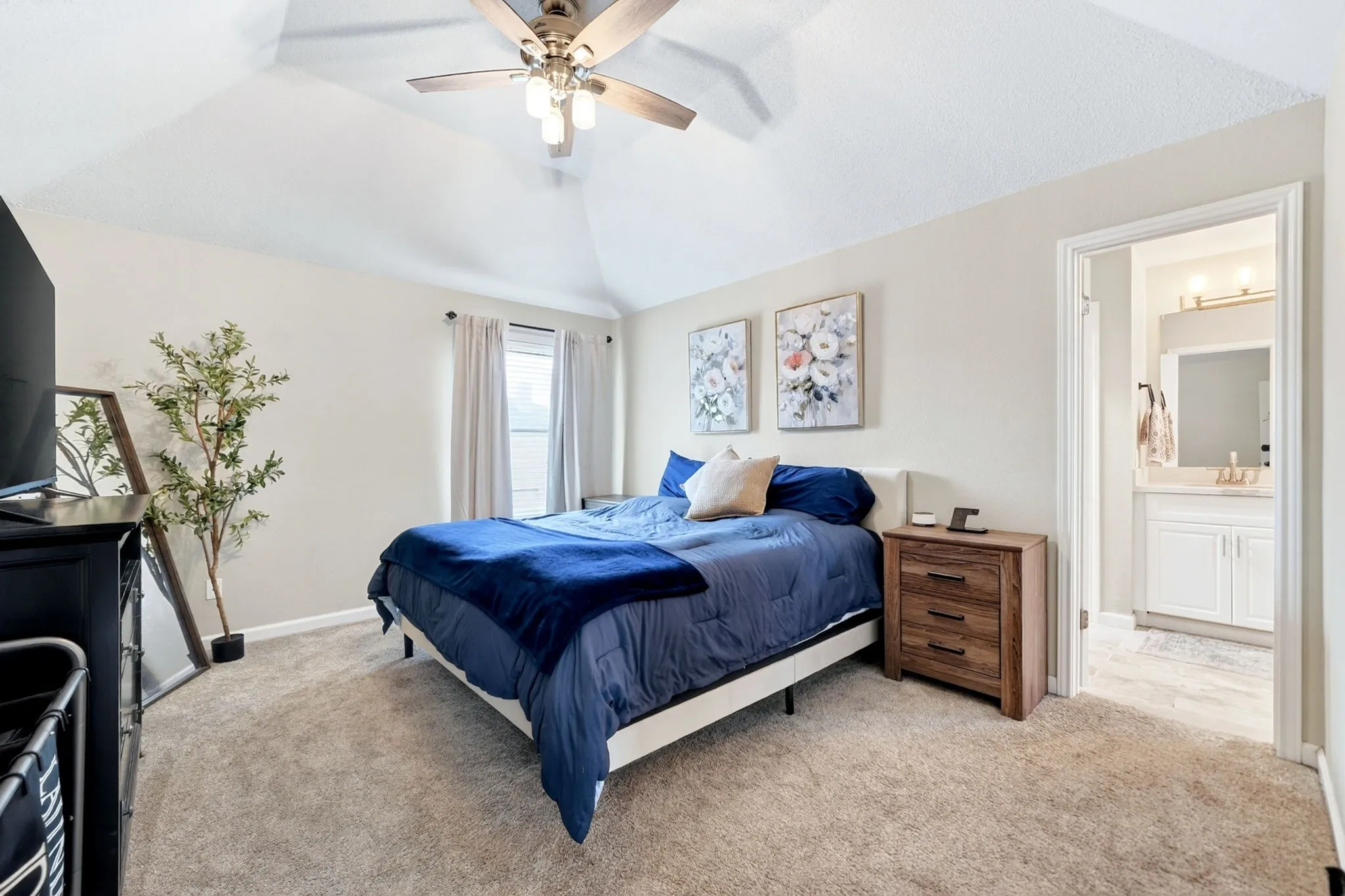 Carpeted bedroom featuring a vaulted ceiling with a ceiling fan, a window with curtains, and a doorway leading to a bathroom with a single vanity and a wall-mounted light fixture