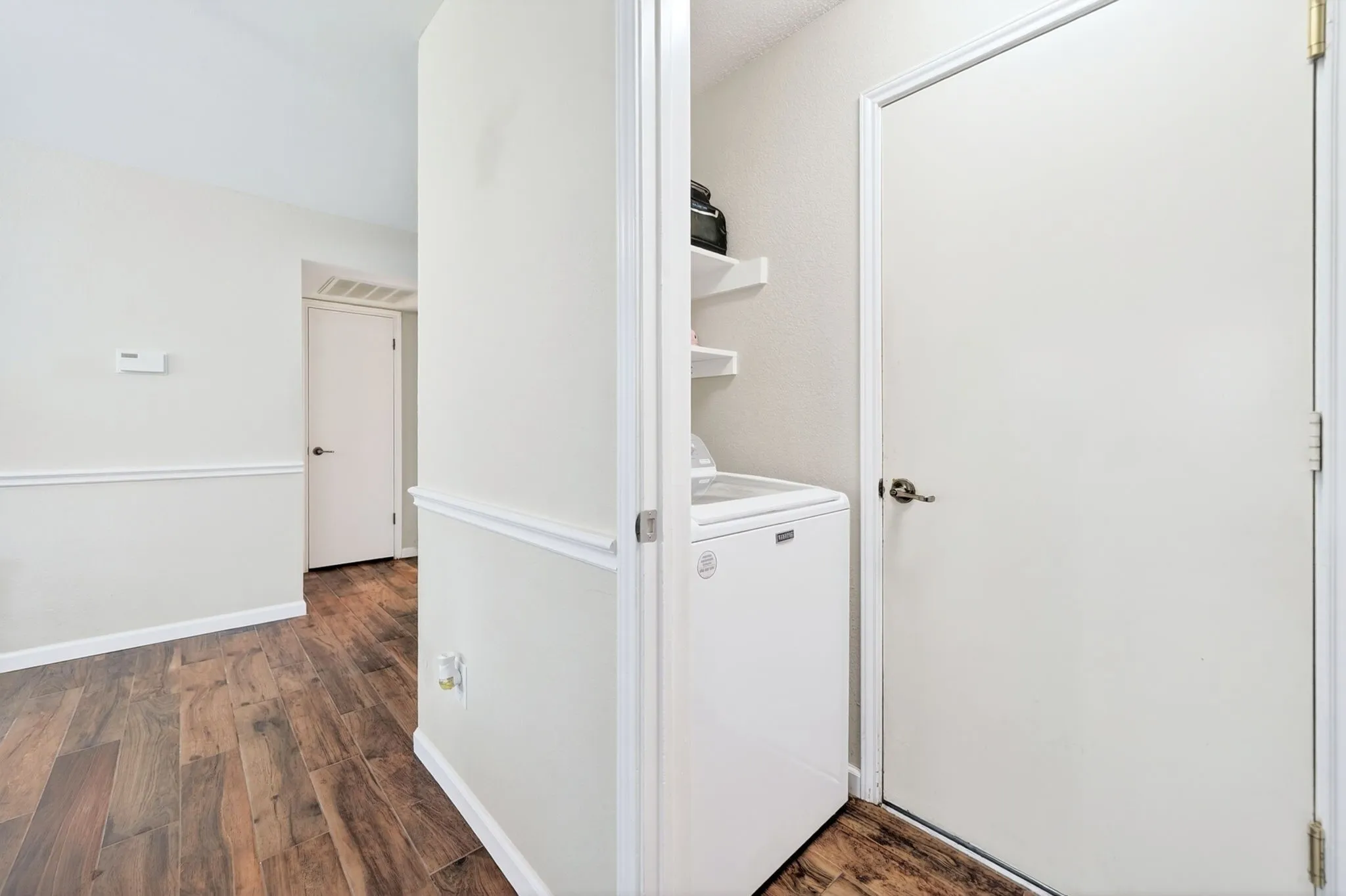 Laundry area featuring a top-load washing machine, white floating shelves, and wood-finish flooring