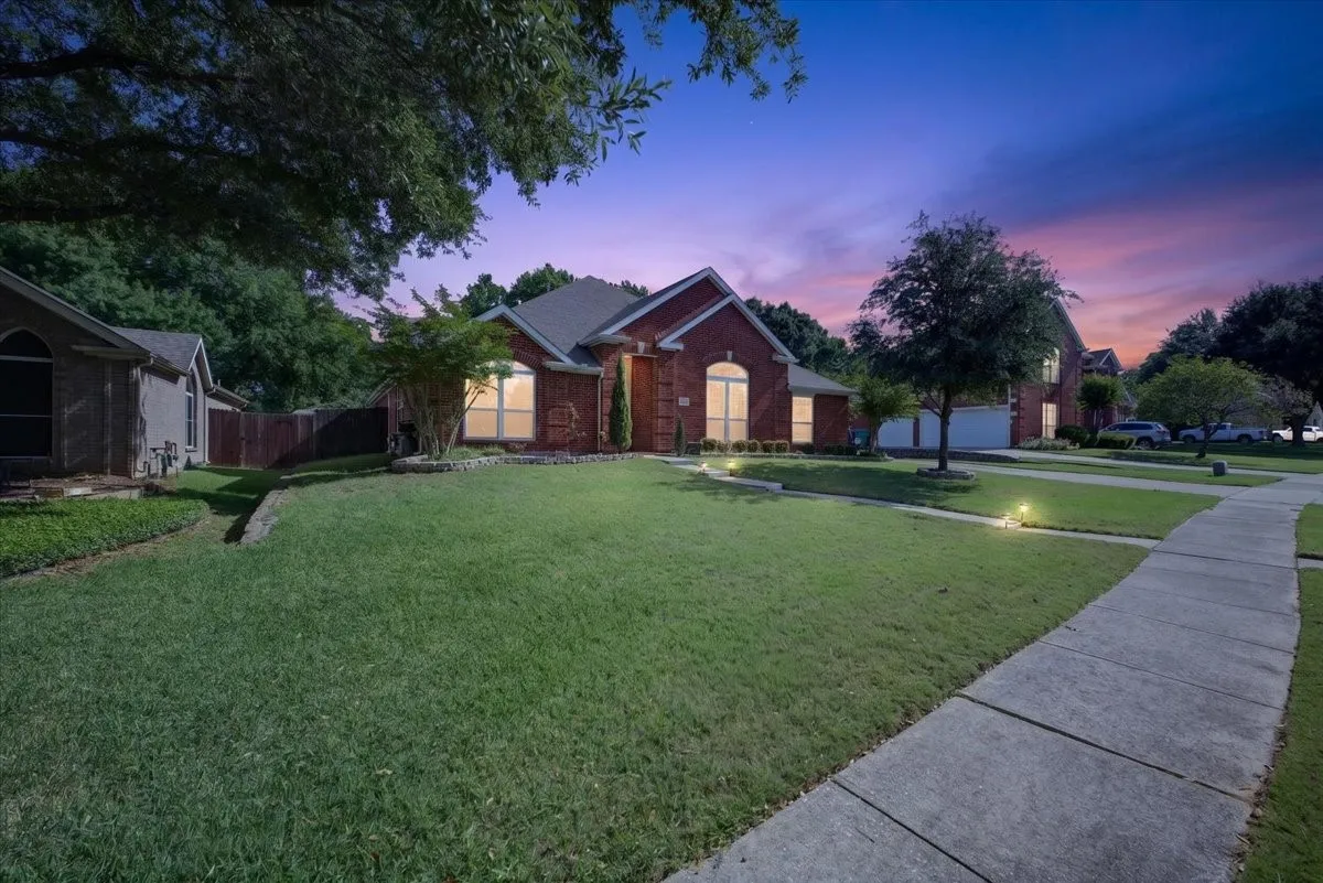 Front exterior featuring a brick facade, landscaped lawn, mature trees, and integrated ground lighting