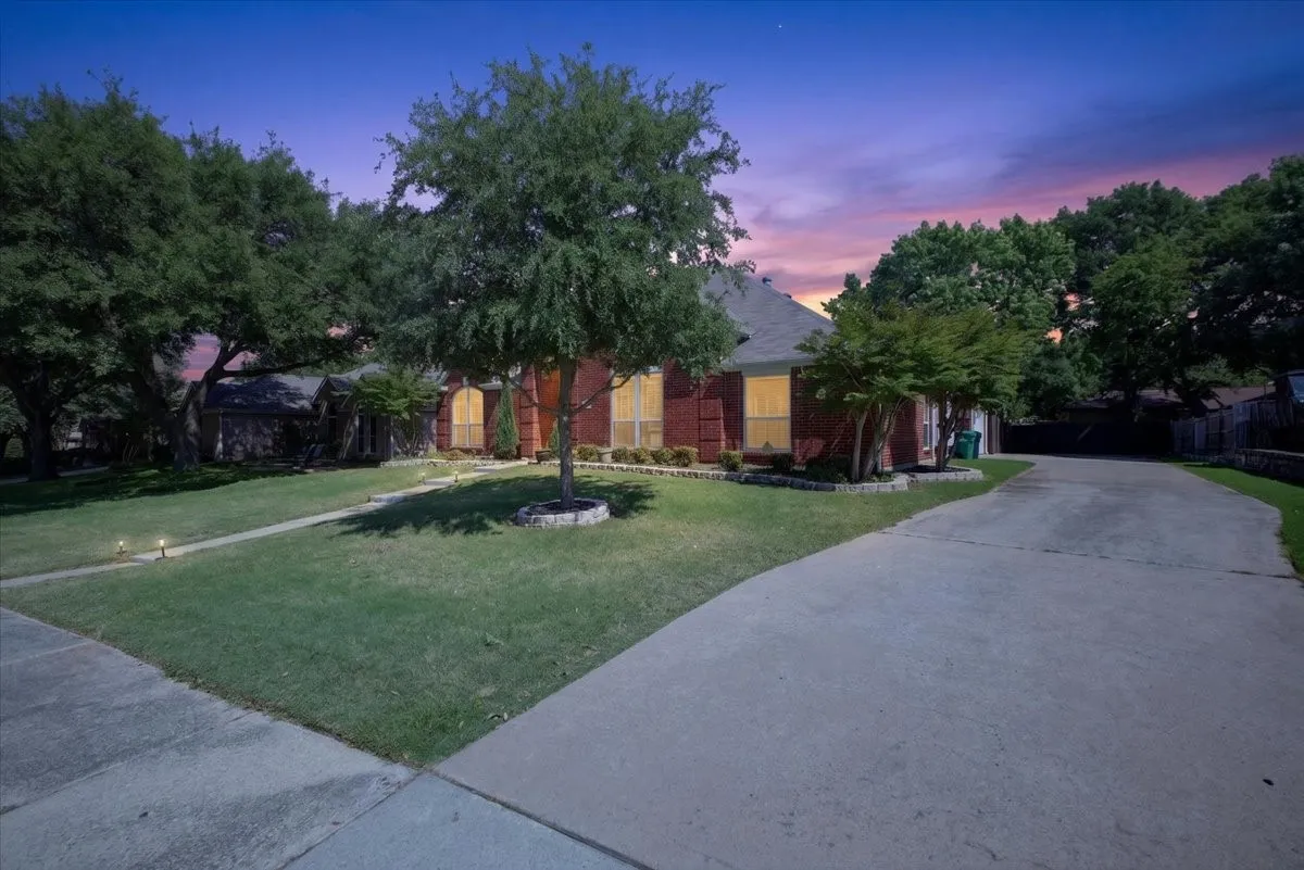 Brick exterior property featuring a pitched roof, front lawn, mature trees, and a concrete driveway