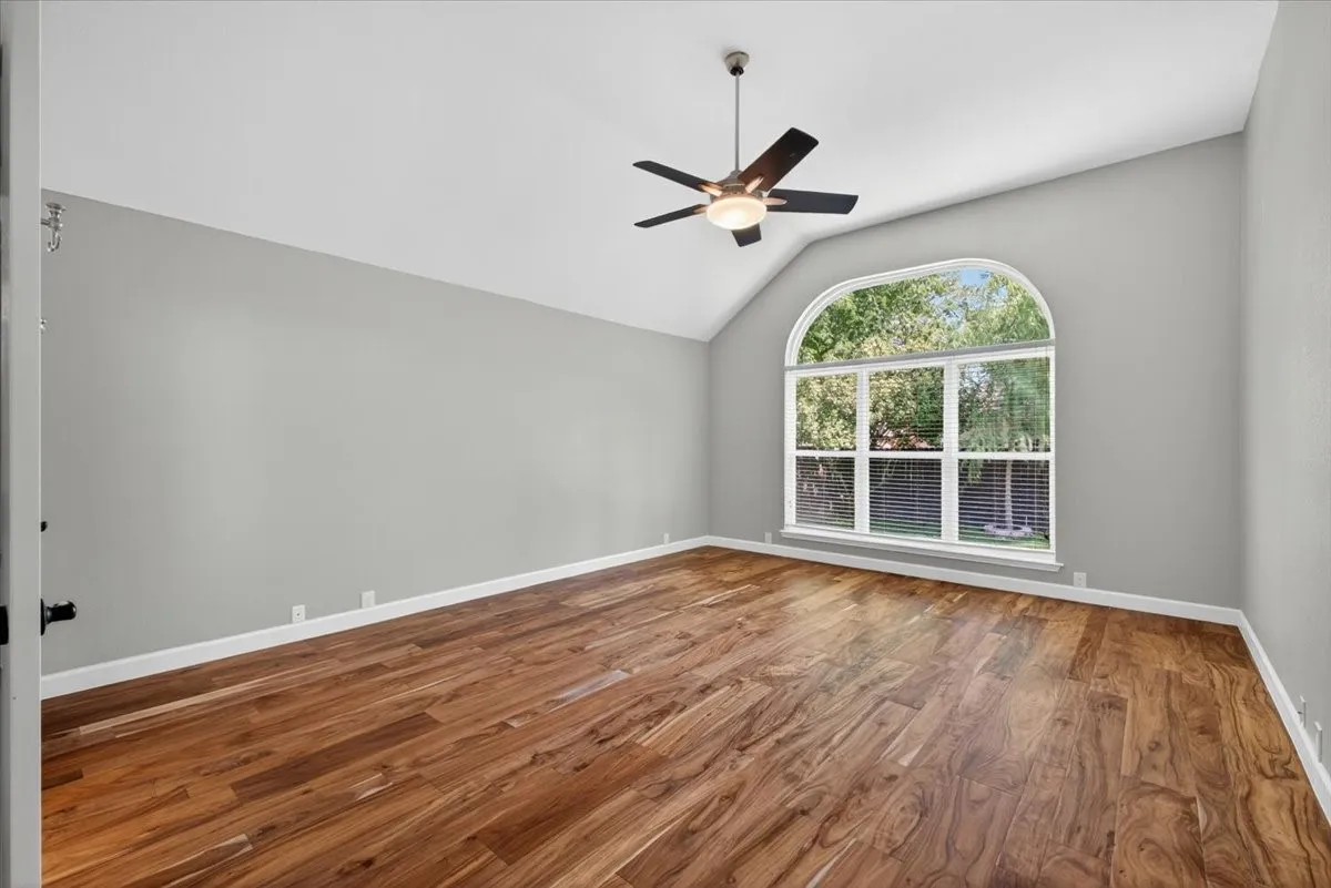 Vaulted ceiling room featuring wood-finish flooring, a large arched window, a ceiling fan with integrated lighting, and neutral wall tones