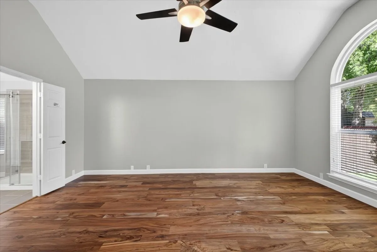 Spacious room featuring vaulted ceilings, wood-finish flooring, a contemporary ceiling fan, and an arched window with horizontal blinds