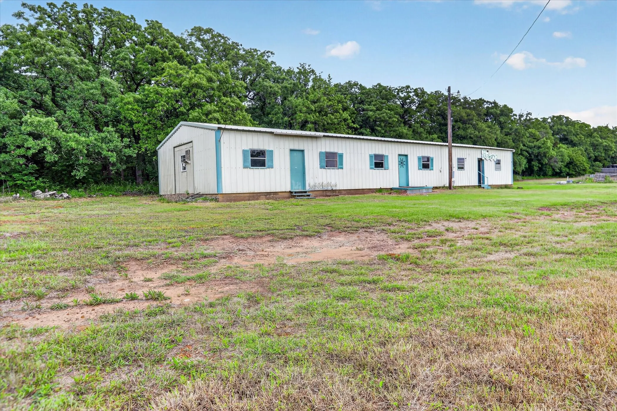 Long white metal structure featuring multiple teal-trimmed windows and doors, set on a green grassy expanse