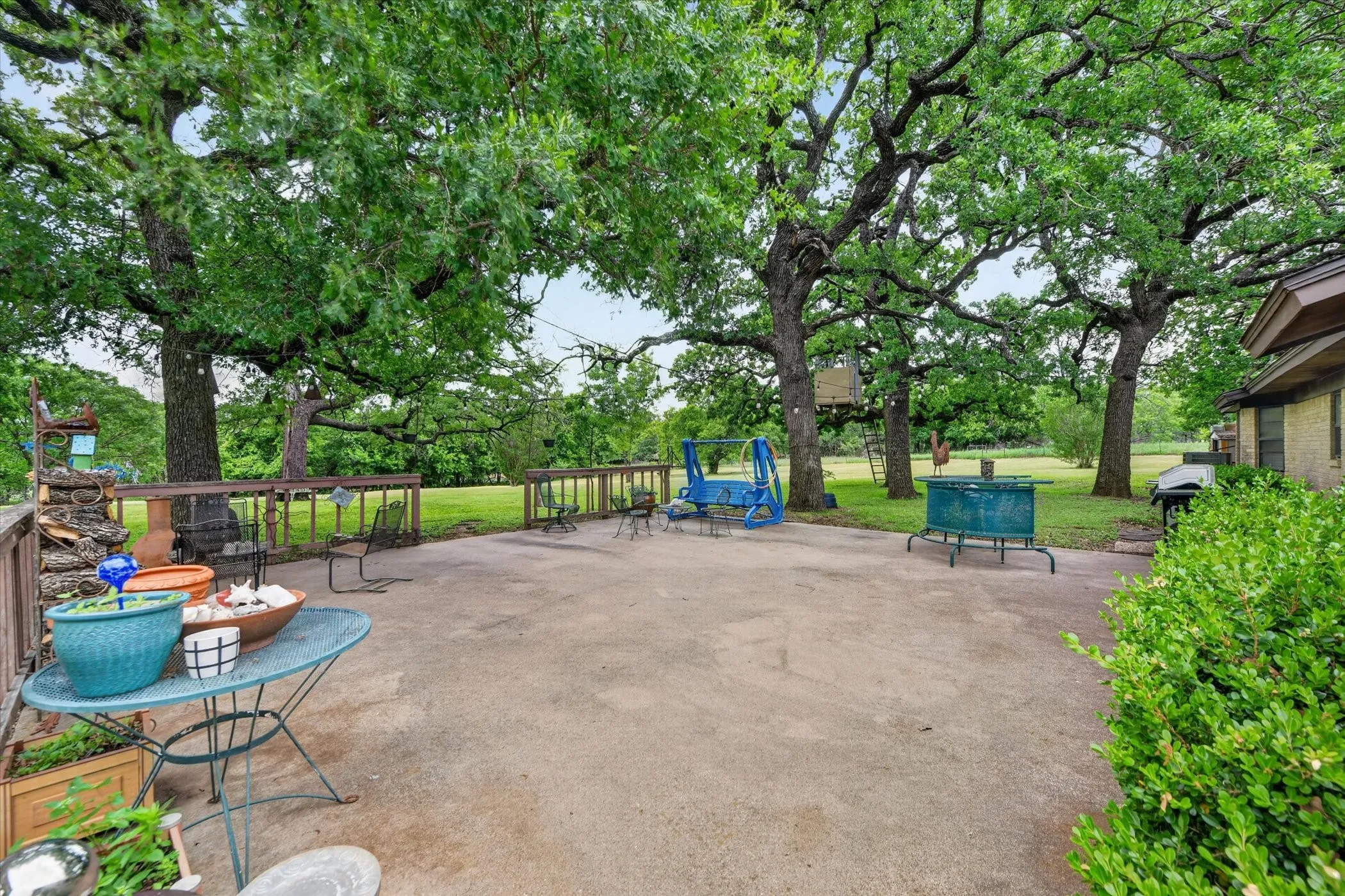 Expansive concrete patio bordered by a wooden deck with railings, mature trees, and a manicured lawn