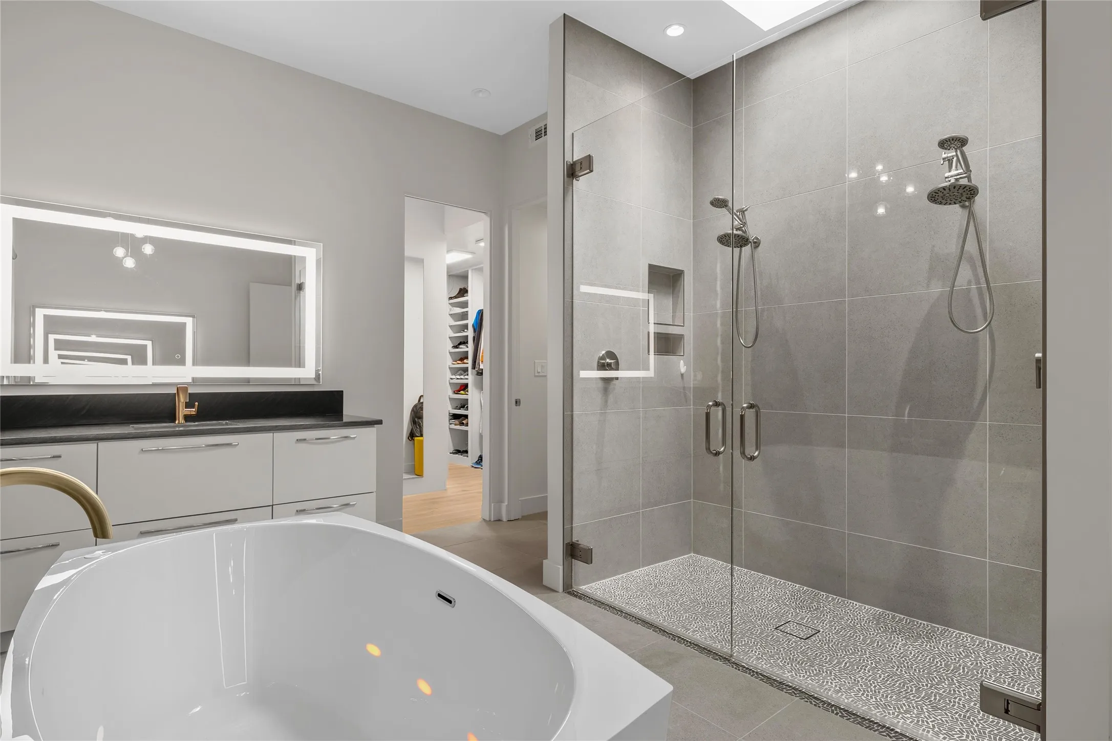 Contemporary bathroom featuring a freestanding light-therapy soaking tub, 2 shower heads glass-enclosed shower with pebble flooring, 2 vanities with Quartzite countertops, LED illuminated wall mirrors, chandelier, recessed lighting, his/hers closets