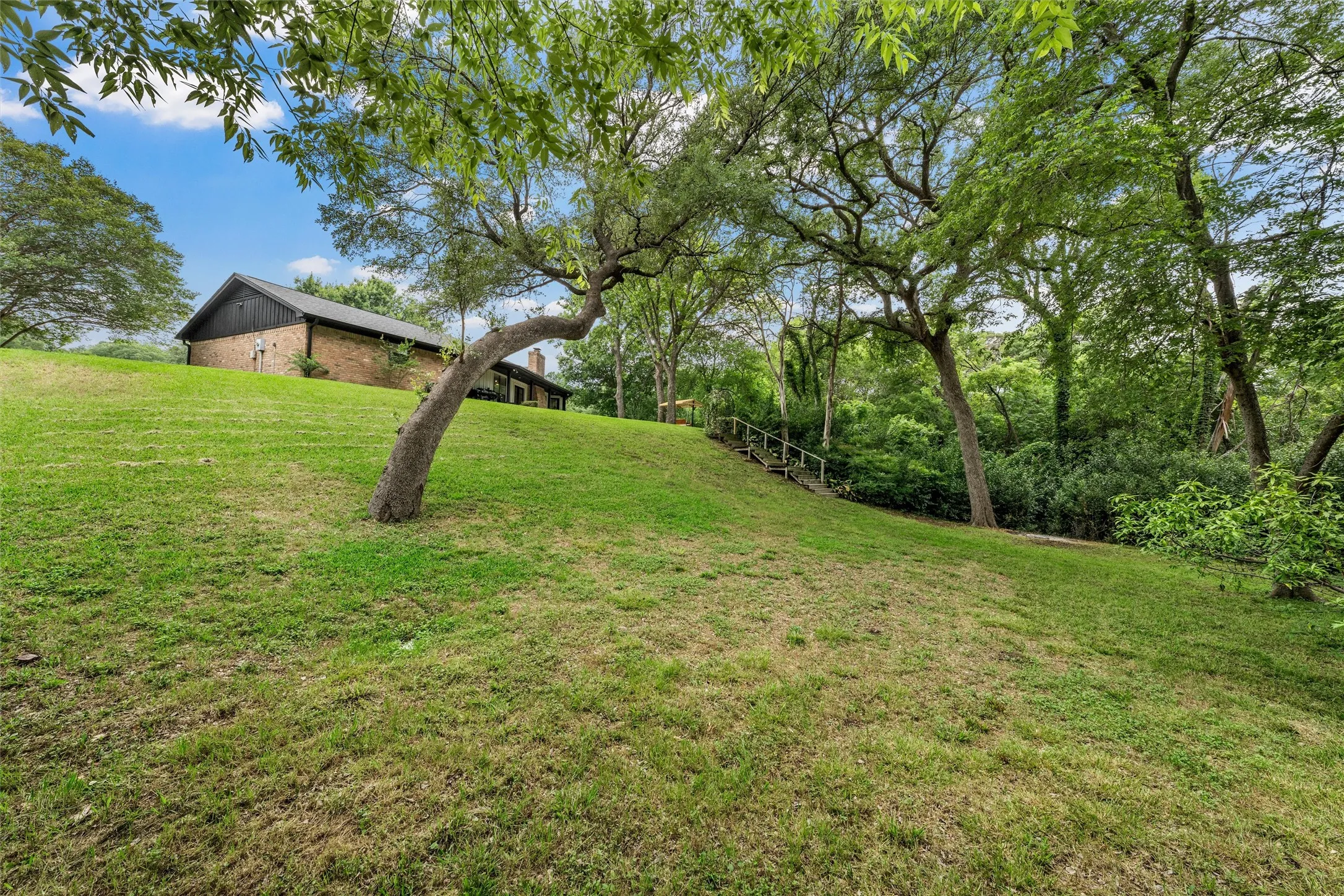 Expansive grassy hillside with mature trees and a brick-sided residence featuring a dark roofline