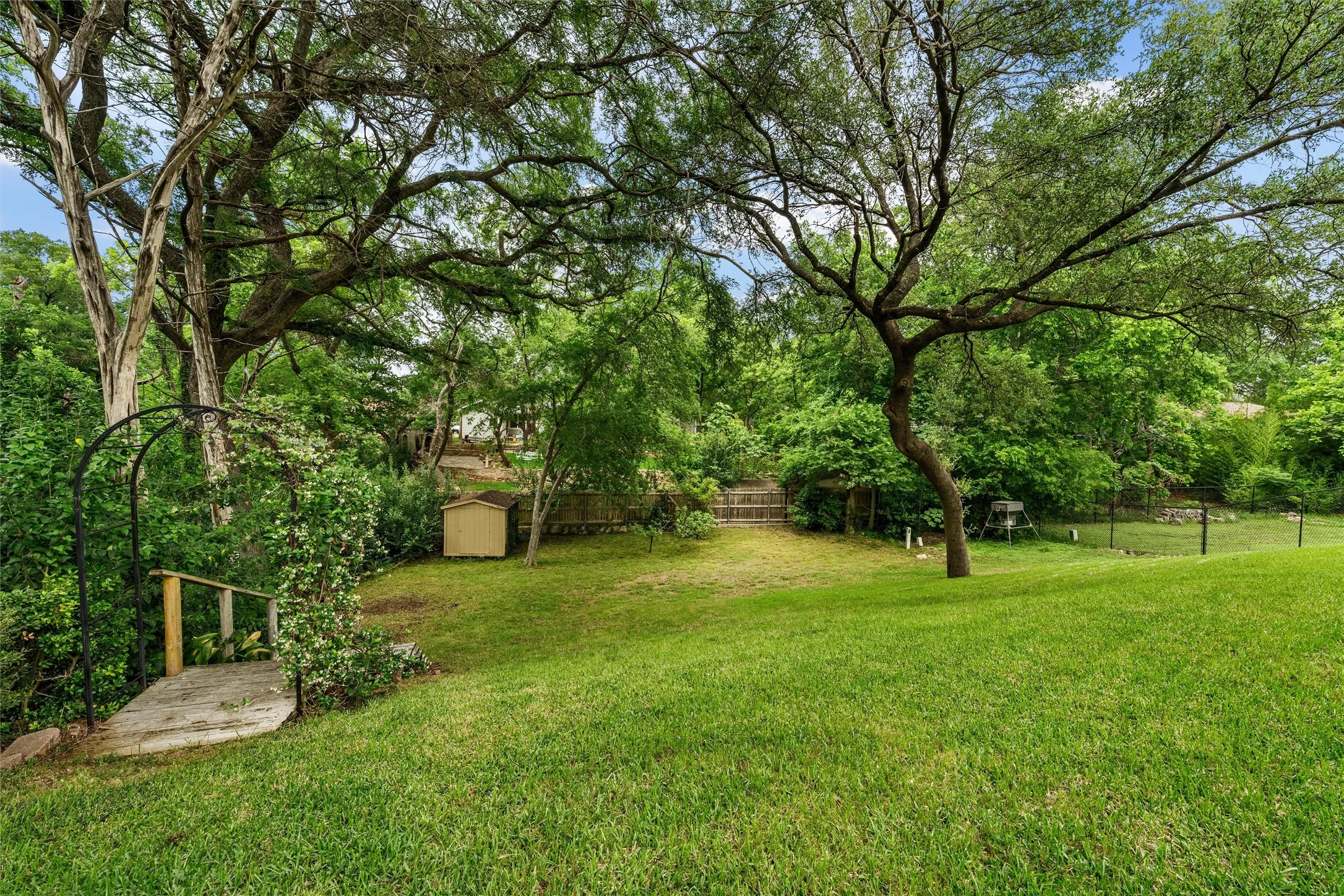 Expansive sloped backyard featuring mature trees, a manicured lawn, a wooden pedestrian bridge with an archway, and a storage shed