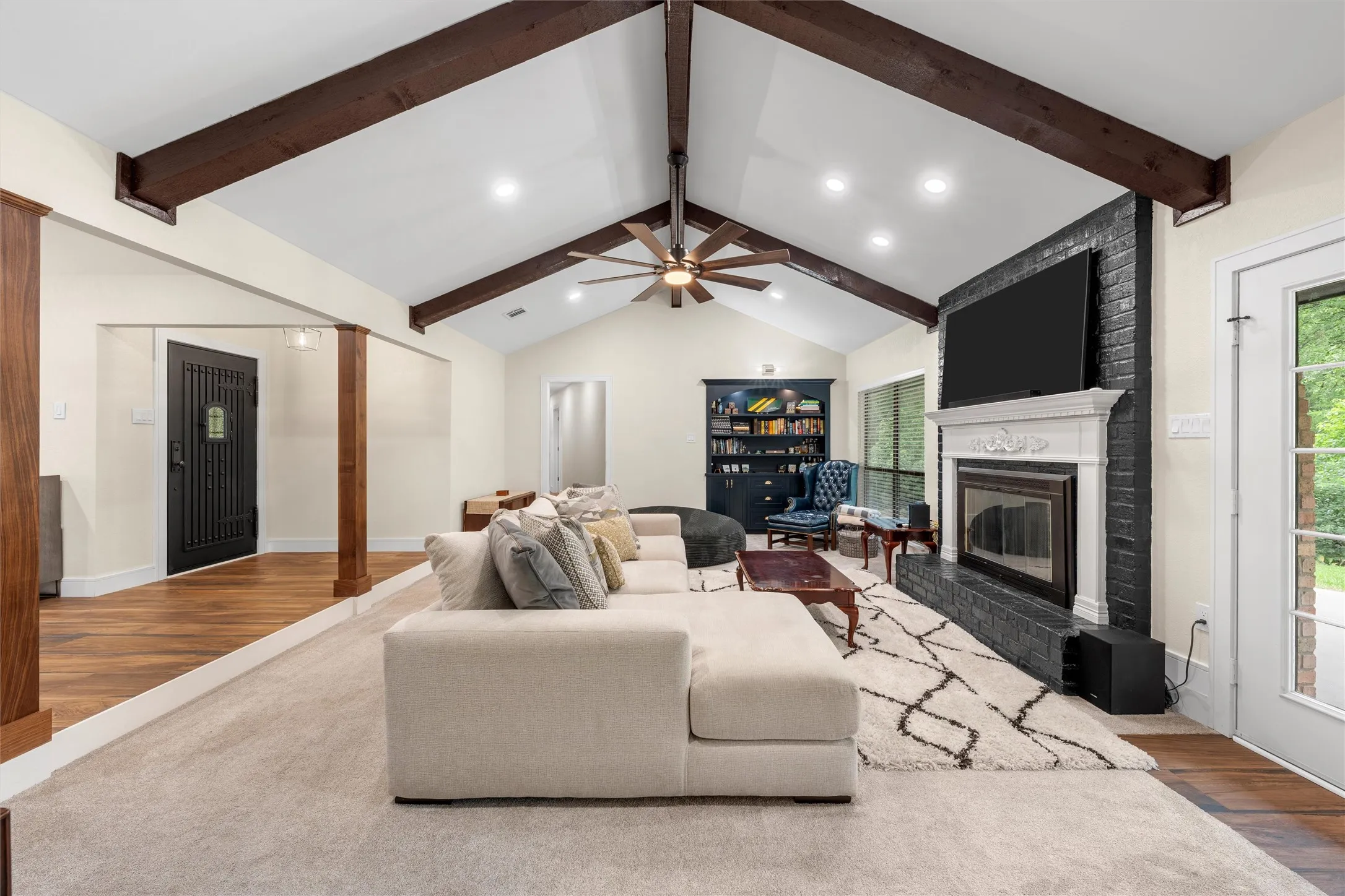 Vaulted ceiling with exposed wood beams, brick fireplace with decorative mantel, wood-finish flooring, recessed lighting, and a ceiling fan