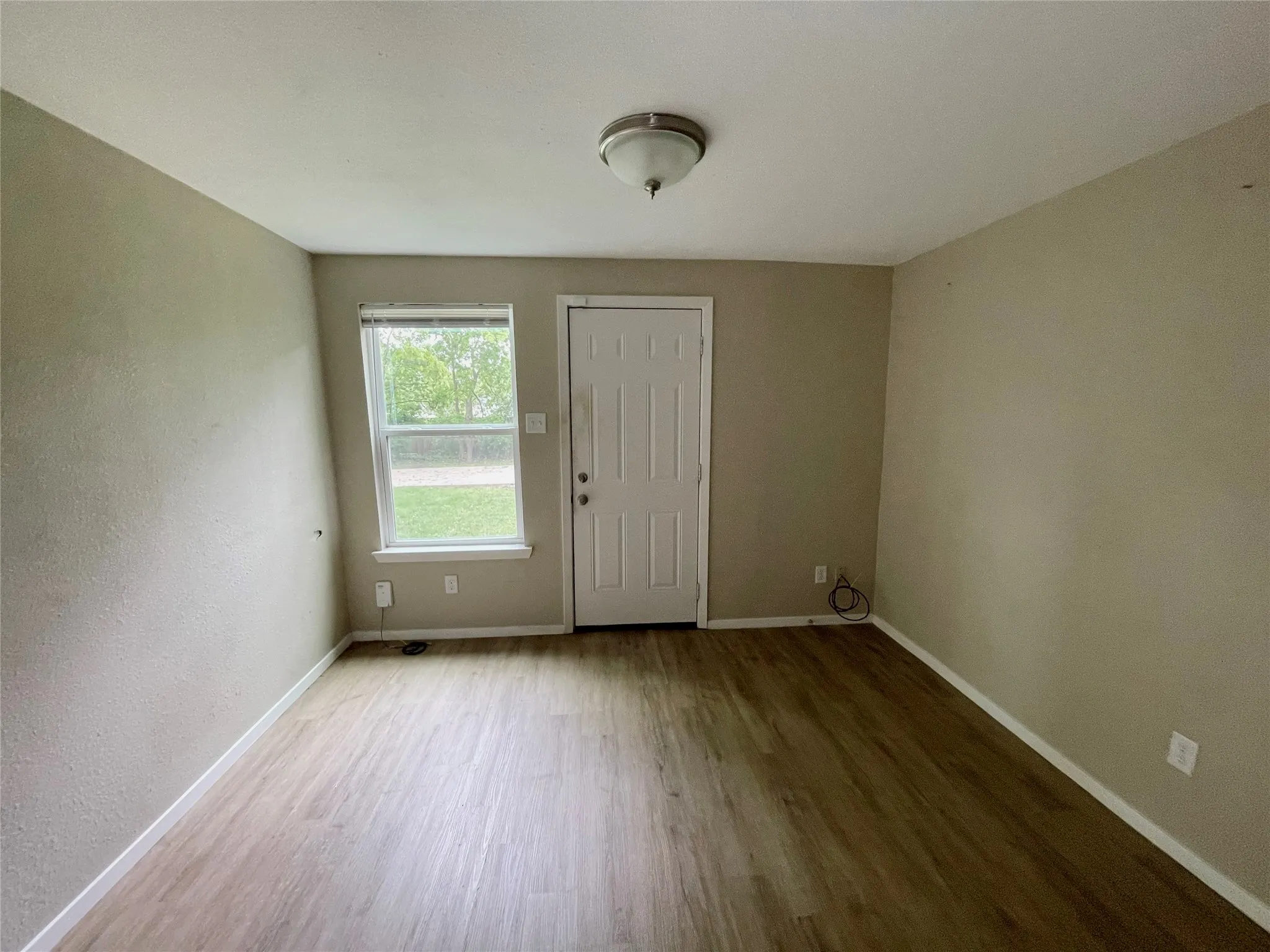 Spacious room featuring wood-finish flooring, neutral-toned walls, a white paneled door, and a single window with blinds