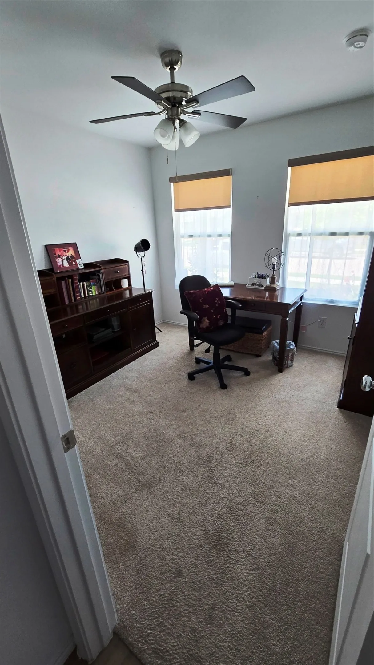 Carpeted interior room featuring two windows with roller shades, a ceiling fan with integrated lighting, and white walls