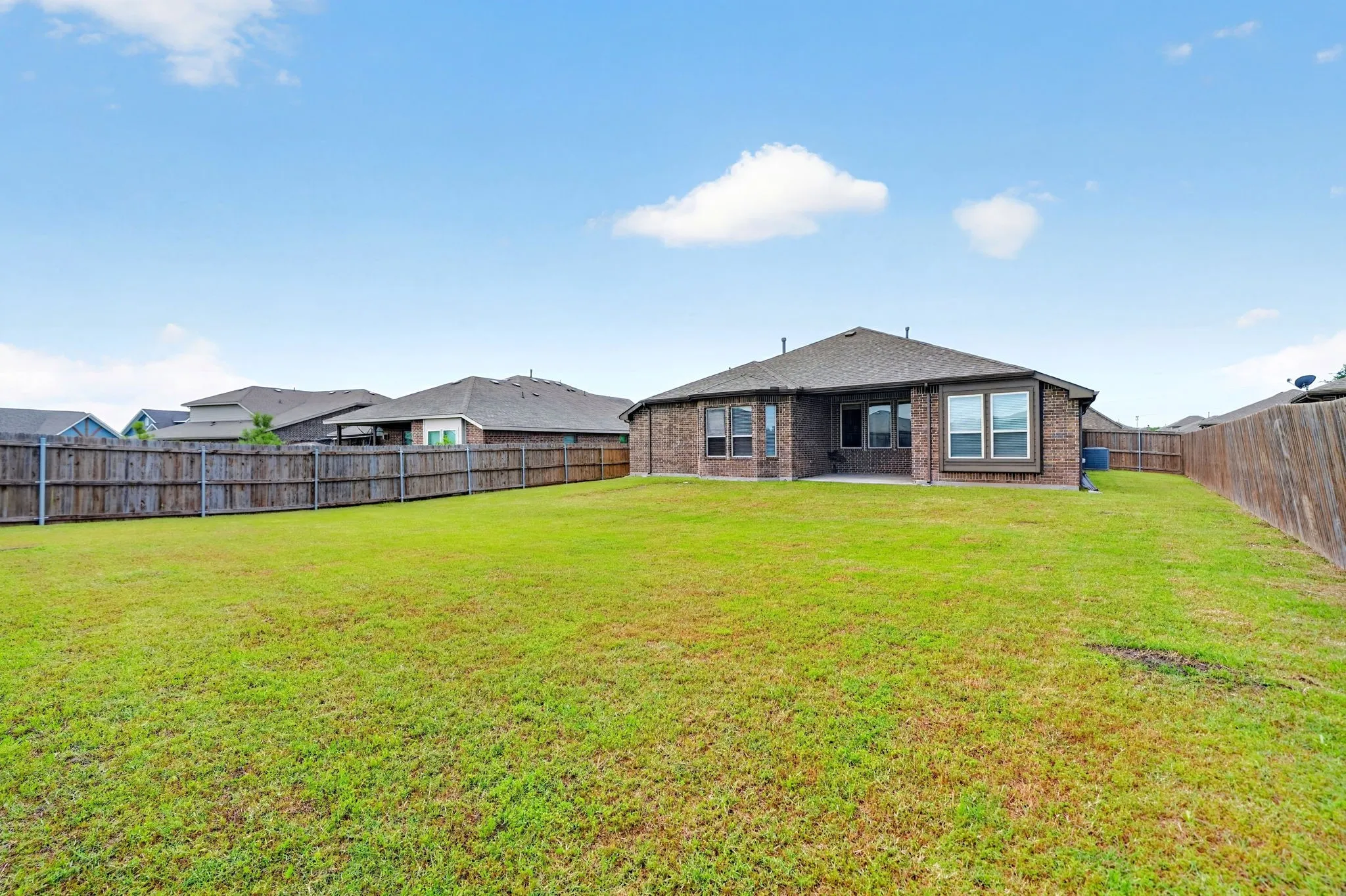 Spacious backyard featuring a large green lawn, full privacy fencing, and a brick exterior home with multiple windows and a covered patio