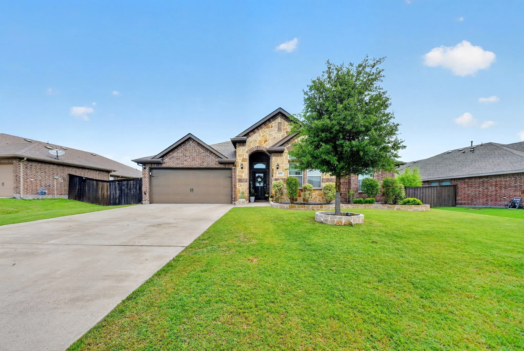 Brick and stone facade residence featuring an arched entryway, attached two-car garage, mature front yard tree with stone surround, and landscaped garden beds